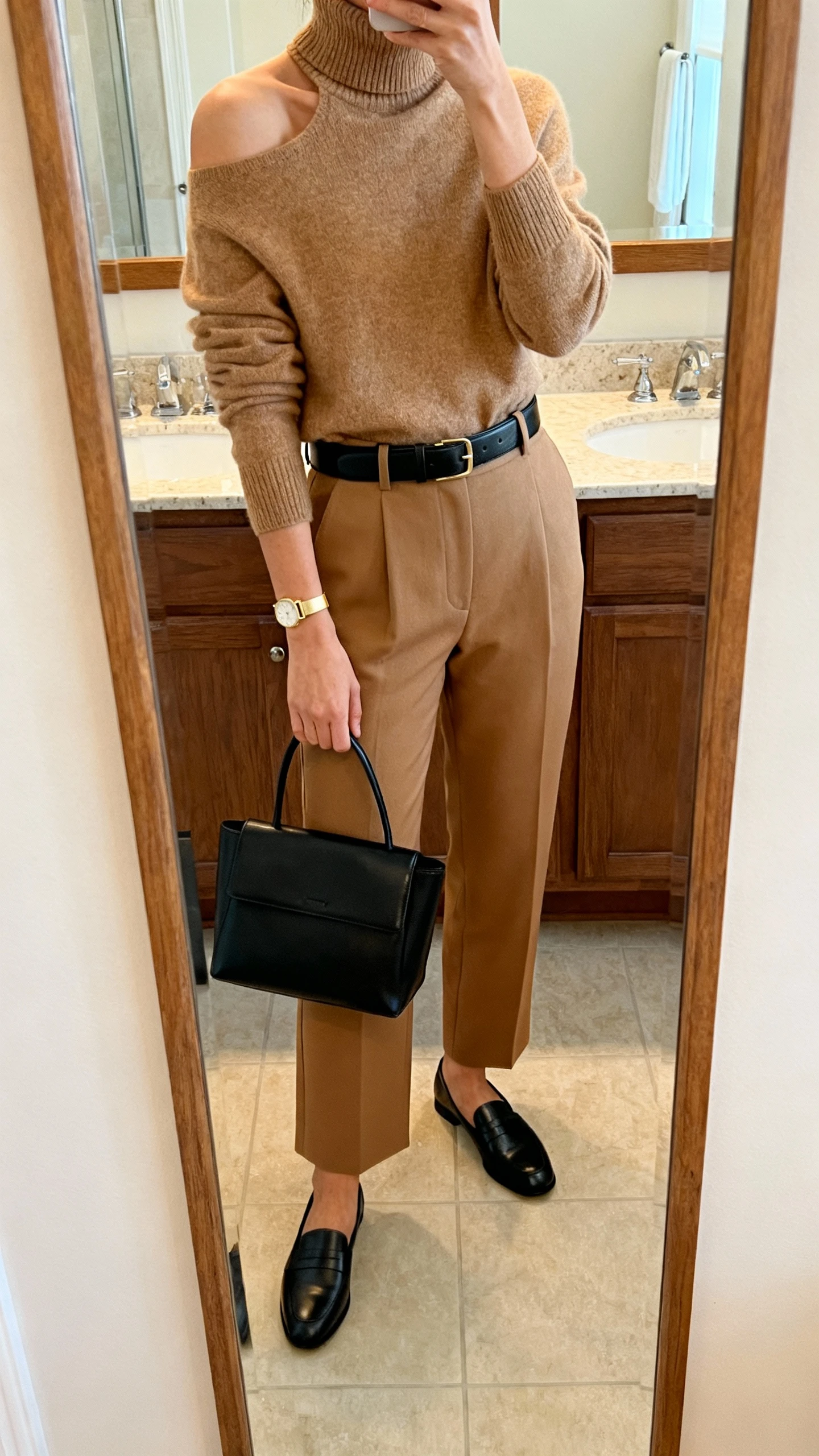 iPhone mirror selfie of a woman in quiet luxury neutrals: camel cashmere turtleneck, tailored tan trousers, black leather belt, black loafers, black structured tote, thin gold watch; face not visible, shoulder-crop, bathroom full-length mirror, warm indoor lighting, iPhone photo quality.