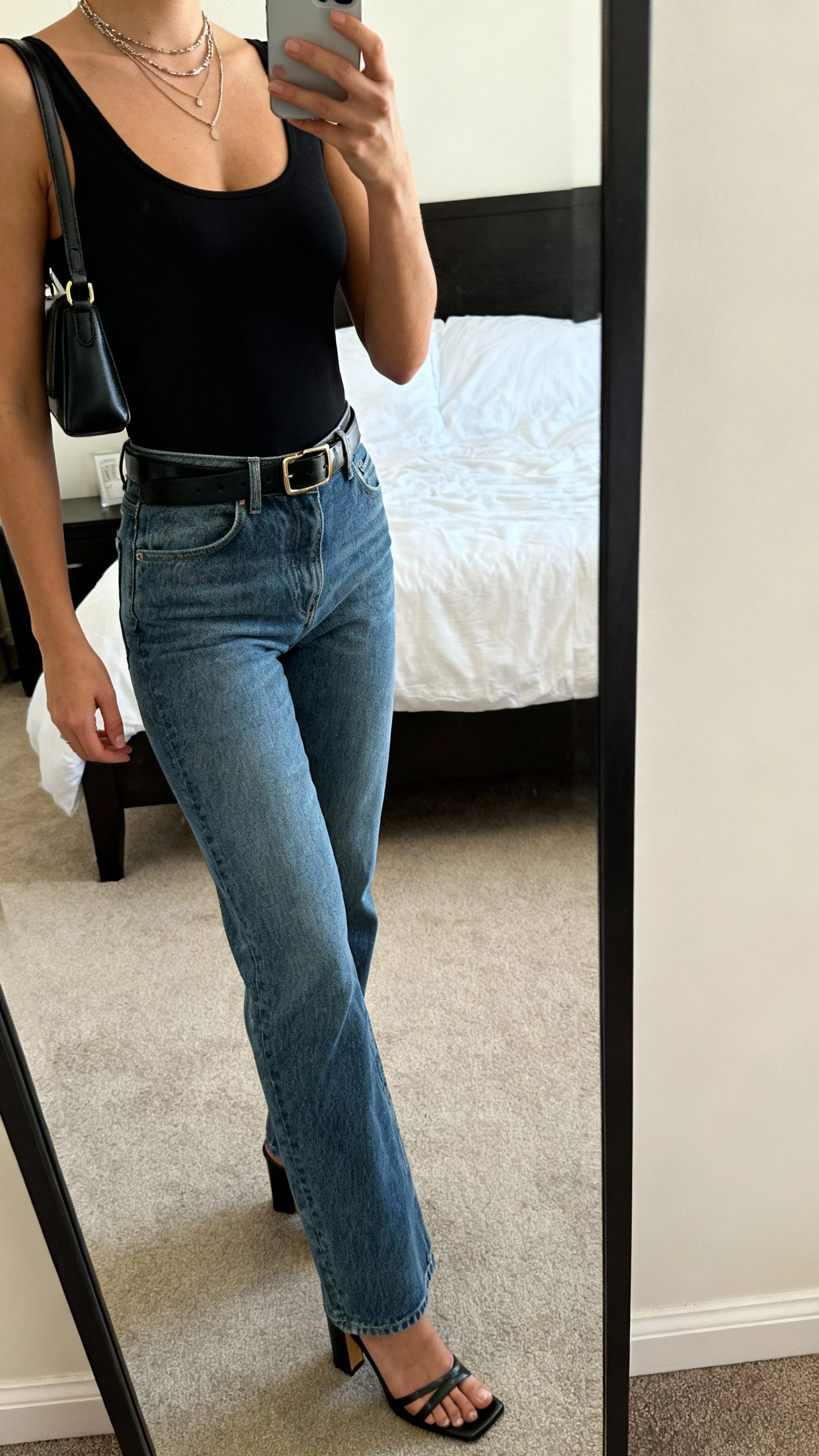 iPhone mirror selfie of a woman in straight-leg mid-wash jeans, sleek black bodysuit, heeled sandals, slim leather belt, small shoulder bag, layered necklaces, face not visible, cropped at shoulders, bedroom mirror, natural daylight, iPhone photo quality.