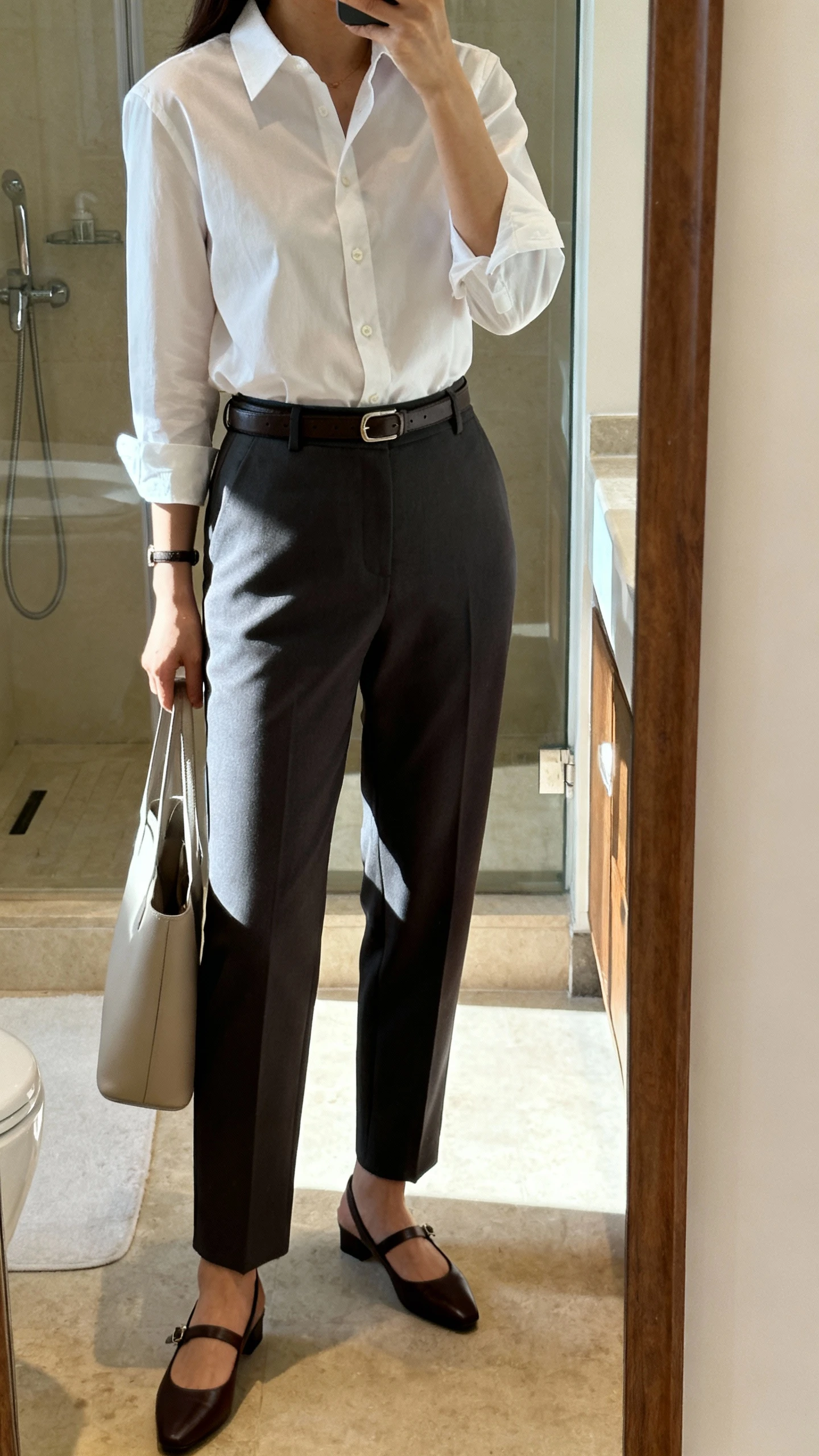 iPhone mirror selfie of a woman in tailored trousers, slingback flats, a tucked-in crisp button-up shirt, slim belt, and minimalist tote, face not visible, side angle in a bathroom full-length mirror, soft indoor morning light, iPhone photo quality.