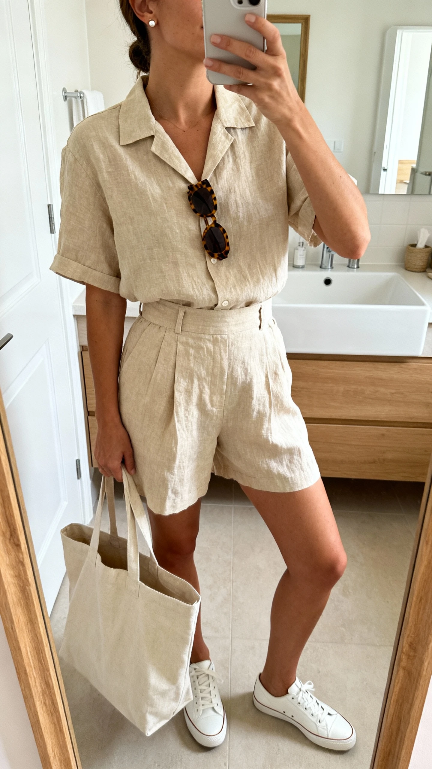iPhone mirror selfie of a woman wearing a beige linen short-sleeve shirt and matching high-waisted linen shorts set, white leather sneakers, oversized canvas tote, tortoiseshell sunglasses tucked on shirt, simple stud earrings, face not visible, bathroom mirror, morning light, iPhone photo quality