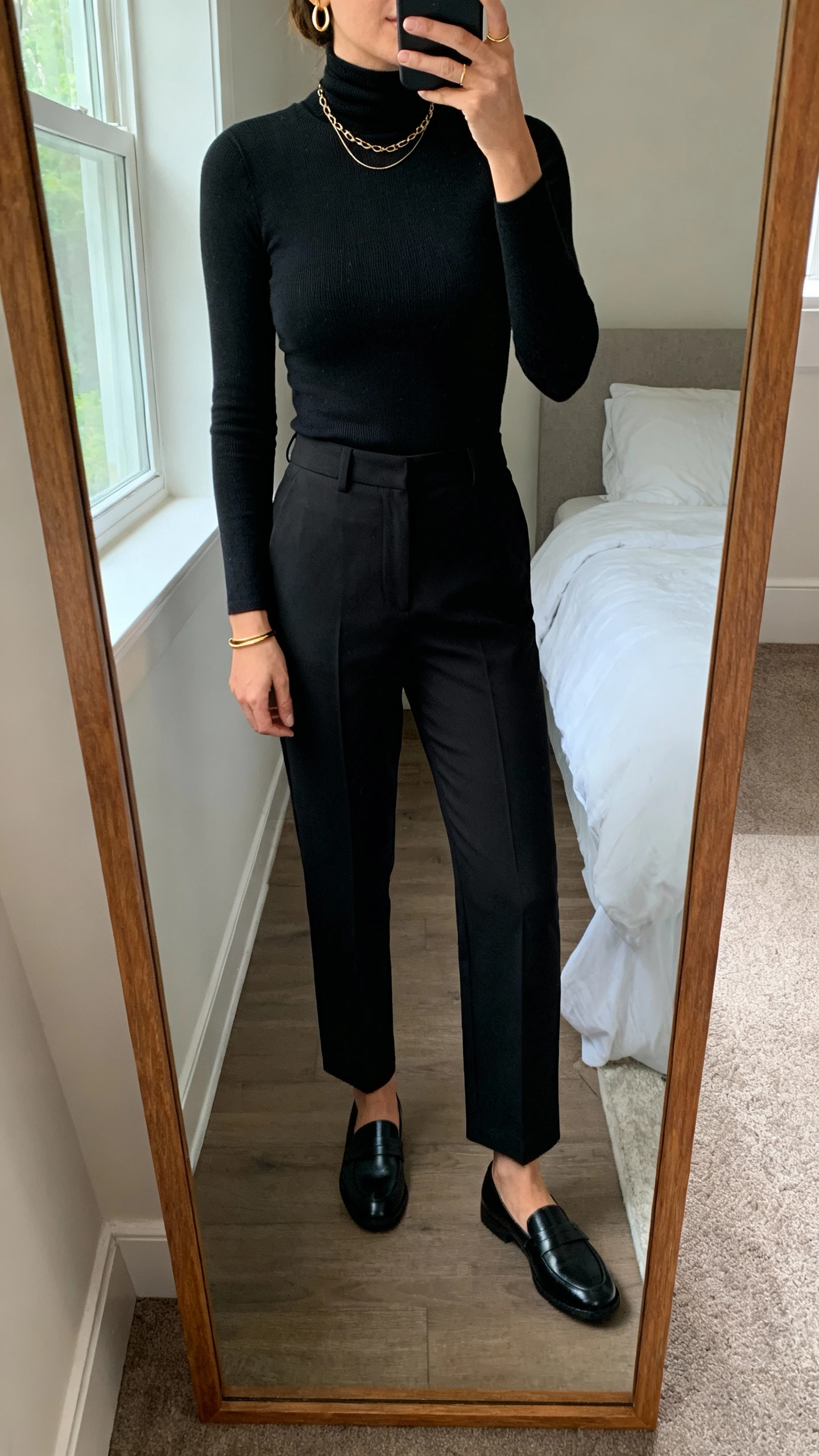 iPhone mirror selfie of a woman wearing a black fitted turtleneck, black tailored straight-leg trousers, sleek black loafers, and minimal gold jewelry (thin hoop earrings, delicate chain necklace, slim bangle), face not visible, cropped at neck, bedroom mirror, natural window light, iPhone photo quality