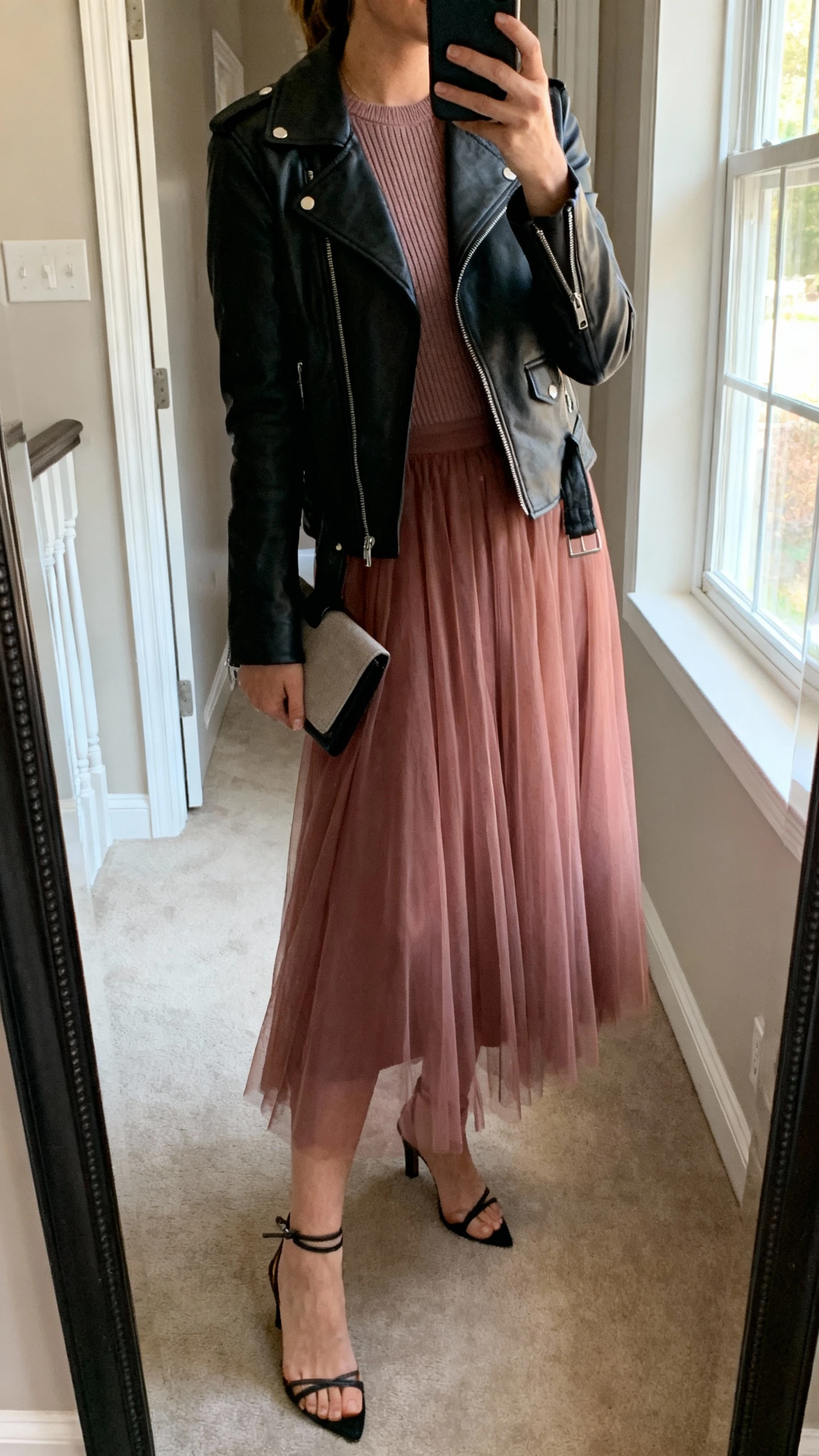iPhone mirror selfie of a woman wearing a black leather biker jacket over a soft tulle midi skirt in dusty rose, fitted knit top, ankle-strap heels, minimalist clutch, face not visible, side angle, entryway full-length mirror, soft afternoon window light, iPhone photo quality.