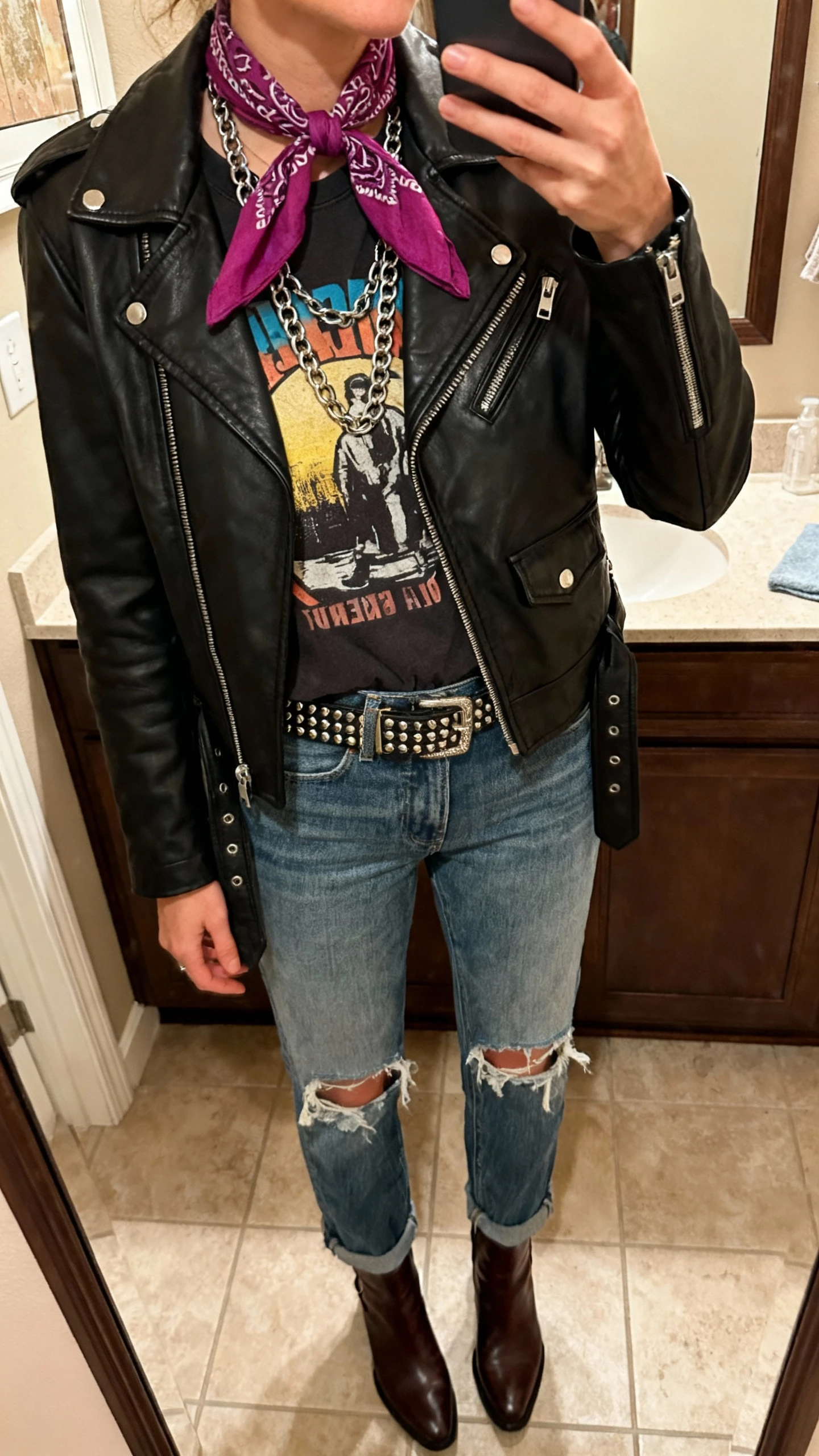 iPhone mirror selfie of a woman wearing a black leather moto jacket, distressed straight-leg denim, graphic tee, ankle boots, a bold saturated scarf tied bandana-style, layered chain necklace, and a studded belt, face not visible, cropped at shoulders, bathroom mirror, warm indoor lighting, iPhone photo quality.