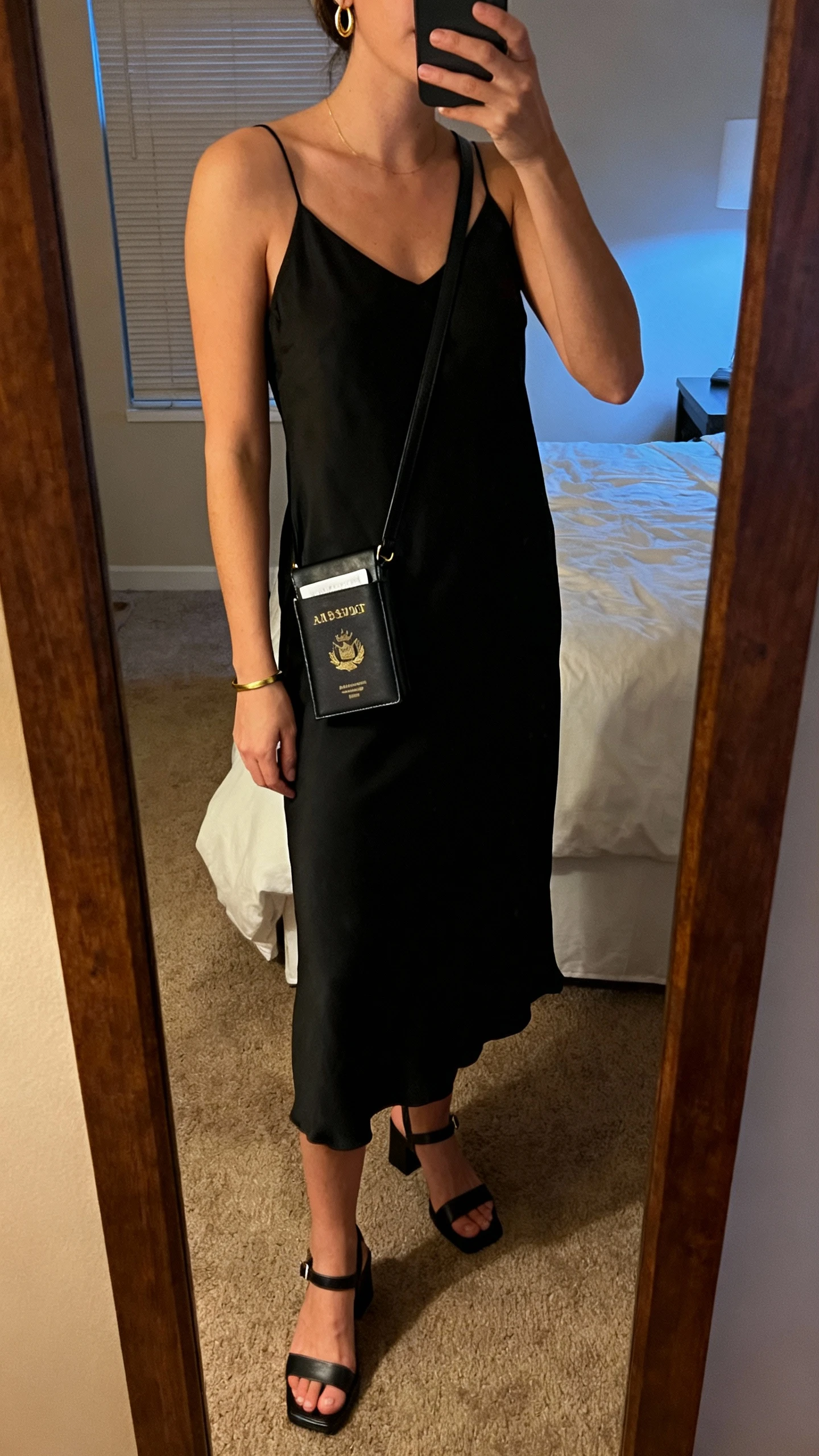 iPhone mirror selfie of a woman wearing a black minimalist slip dress, low block-heel sandals, a slim black leather crossbody that fits a passport, delicate gold hoop earrings, and a thin gold bracelet, face not visible, cropped at shoulders, bedroom full-length mirror, warm evening indoor light, iPhone photo quality