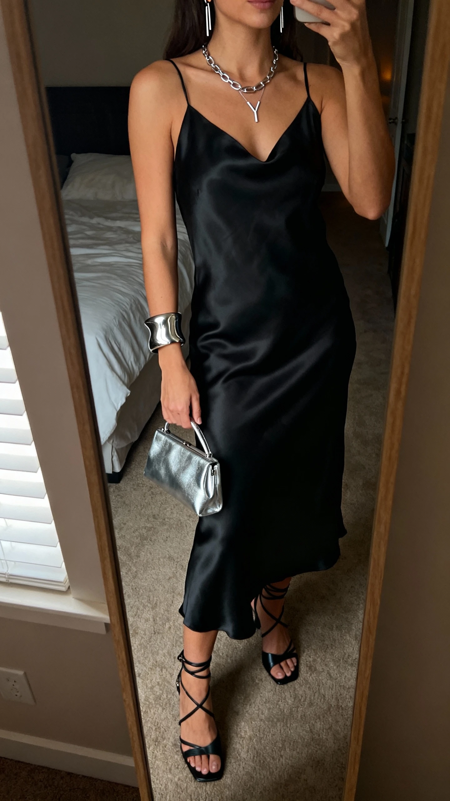 iPhone mirror selfie of a woman wearing a black satin slip dress, strappy black heels, and sleek silver jewelry (Y-chain necklace, threader earrings, sculptural cuff), carrying a small metallic silver clutch, face not visible, cropped at shoulders, bedroom mirror, low evening indoor light, iPhone photo quality
