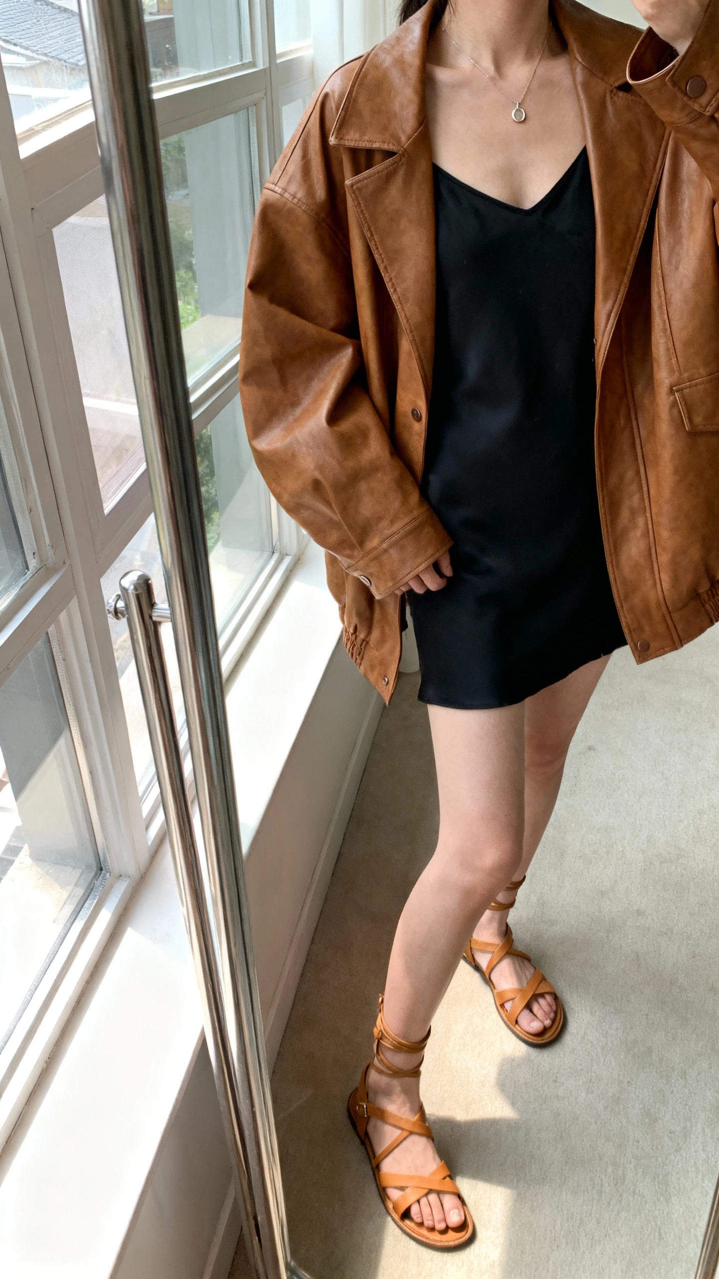 iPhone mirror selfie of a woman wearing a black slip dress layered with an oversized brown leather jacket, delicate pendant necklace, and strappy tan sandals, face not visible, cropped at neck, full-length mirror by window, soft morning light, iPhone photo quality.
