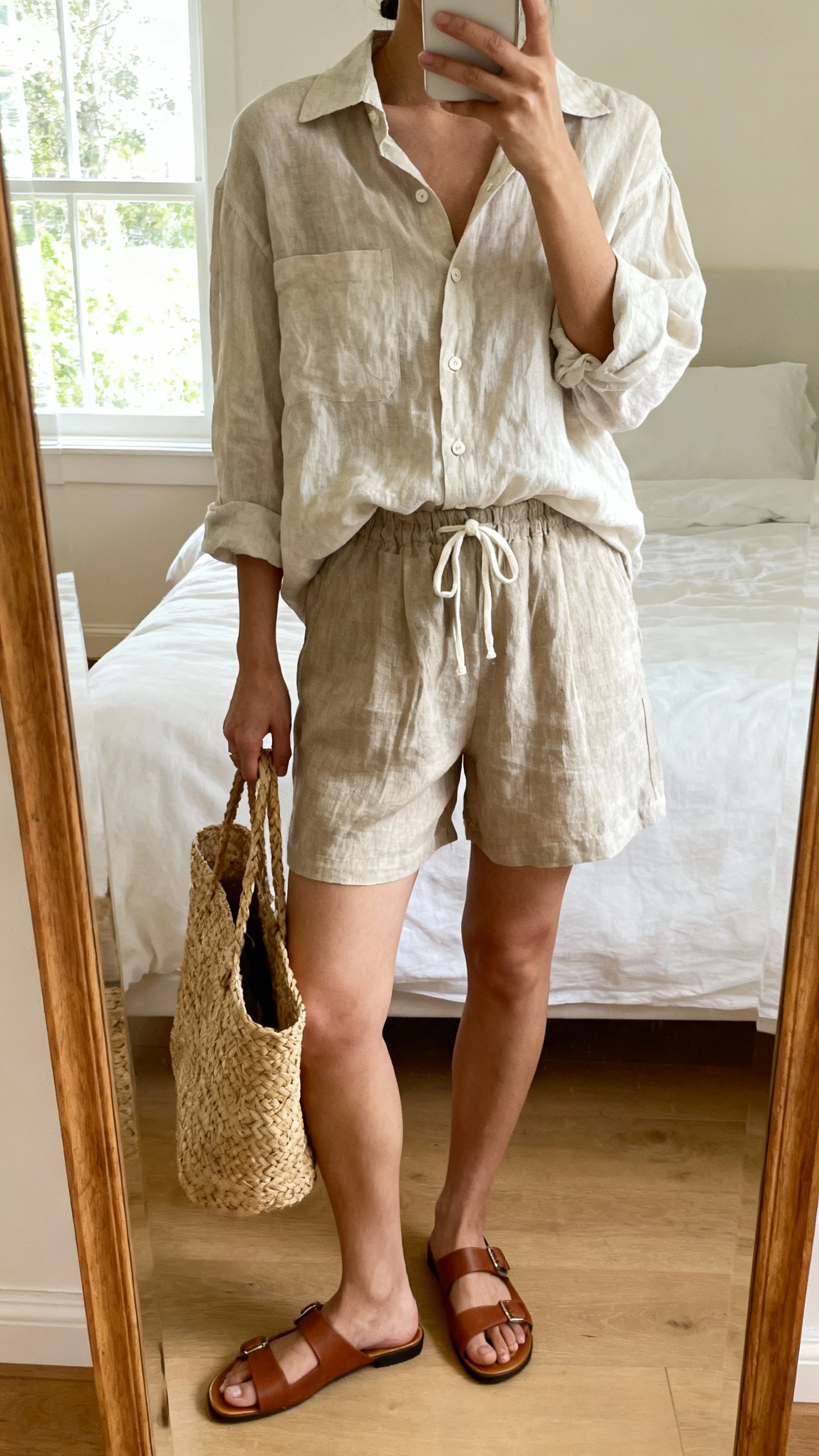 iPhone mirror selfie of a woman wearing a breezy linen matching set (linen button-up shirt and linen drawstring shorts) with a woven tote and flat leather sandals, face not visible, cropped at neck, bedroom mirror, natural window light, iPhone photo quality.