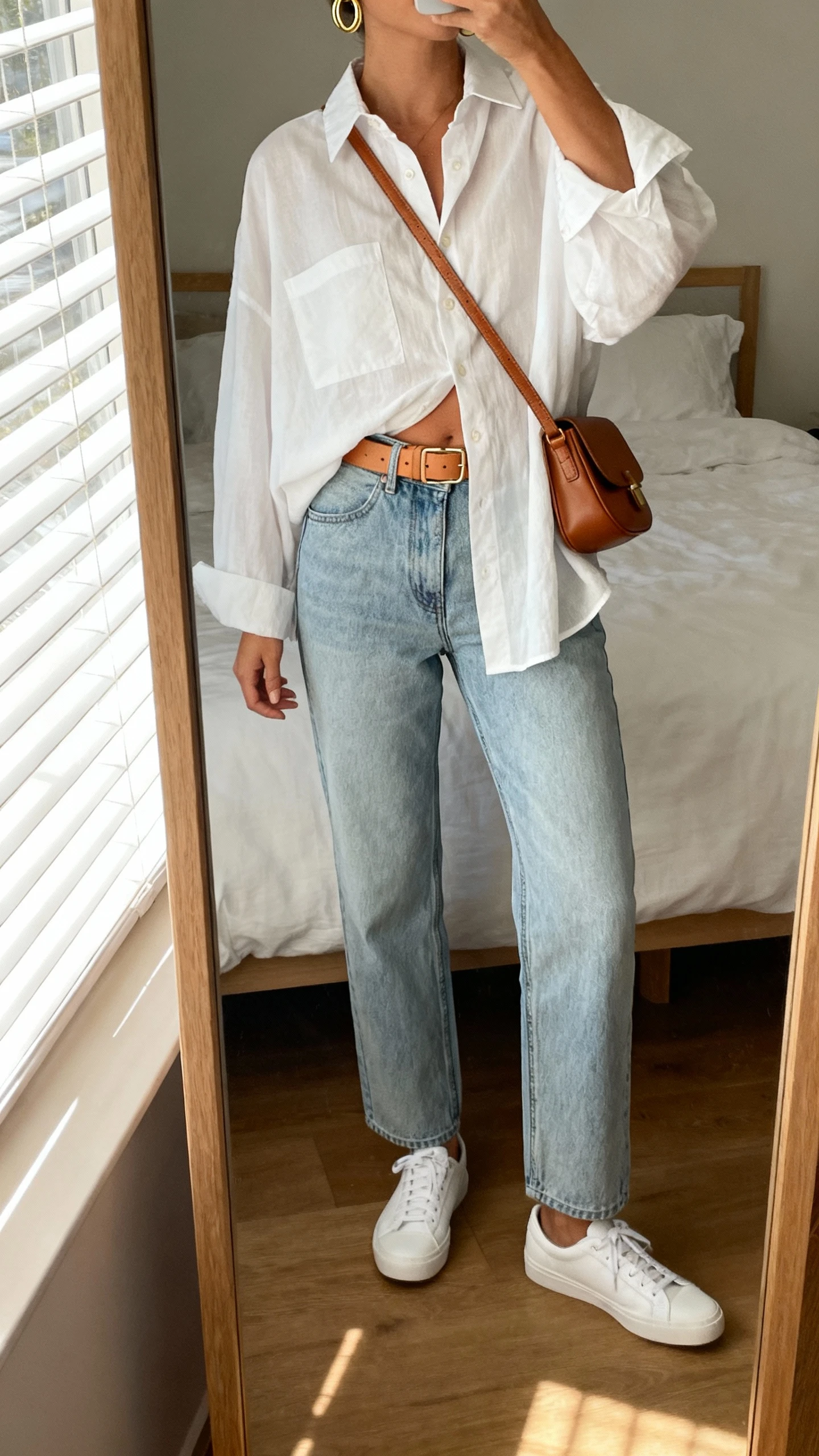 iPhone mirror selfie of a woman wearing a breezy oversized white button-down half-tucked into straight-leg light-wash denim, tan leather belt, simple gold hoops, brown crossbody bag, and white low-top sneakers; face not visible, cropped at neck, bedroom mirror with natural morning window light, iPhone photo quality.