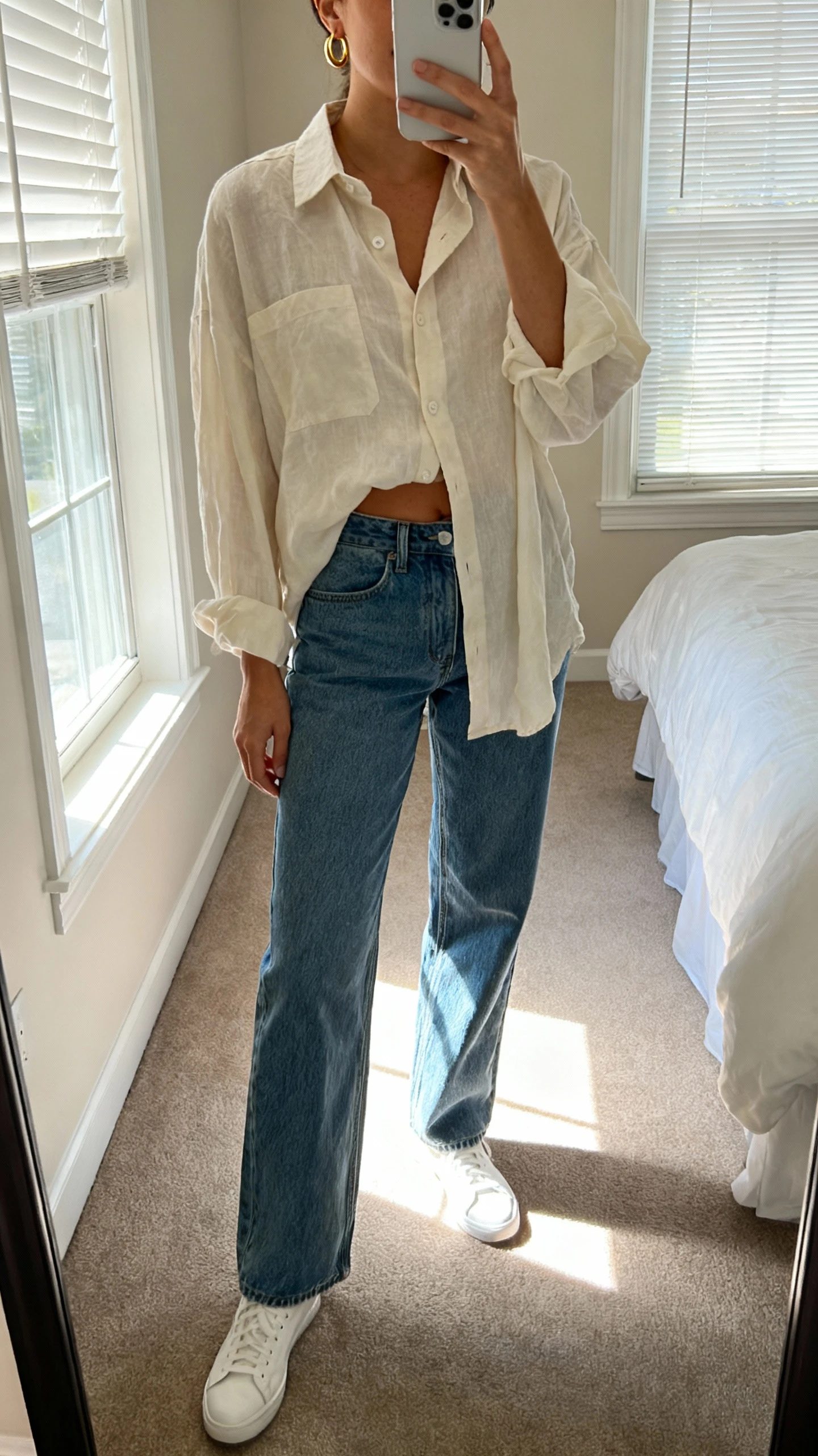 iPhone mirror selfie of a woman wearing a breezy oversized button-down shirt half-tucked into relaxed straight-leg denim with simple white sneakers and minimal gold hoops, face not visible, cropped at neck, bedroom mirror with morning window light, iPhone photo quality.