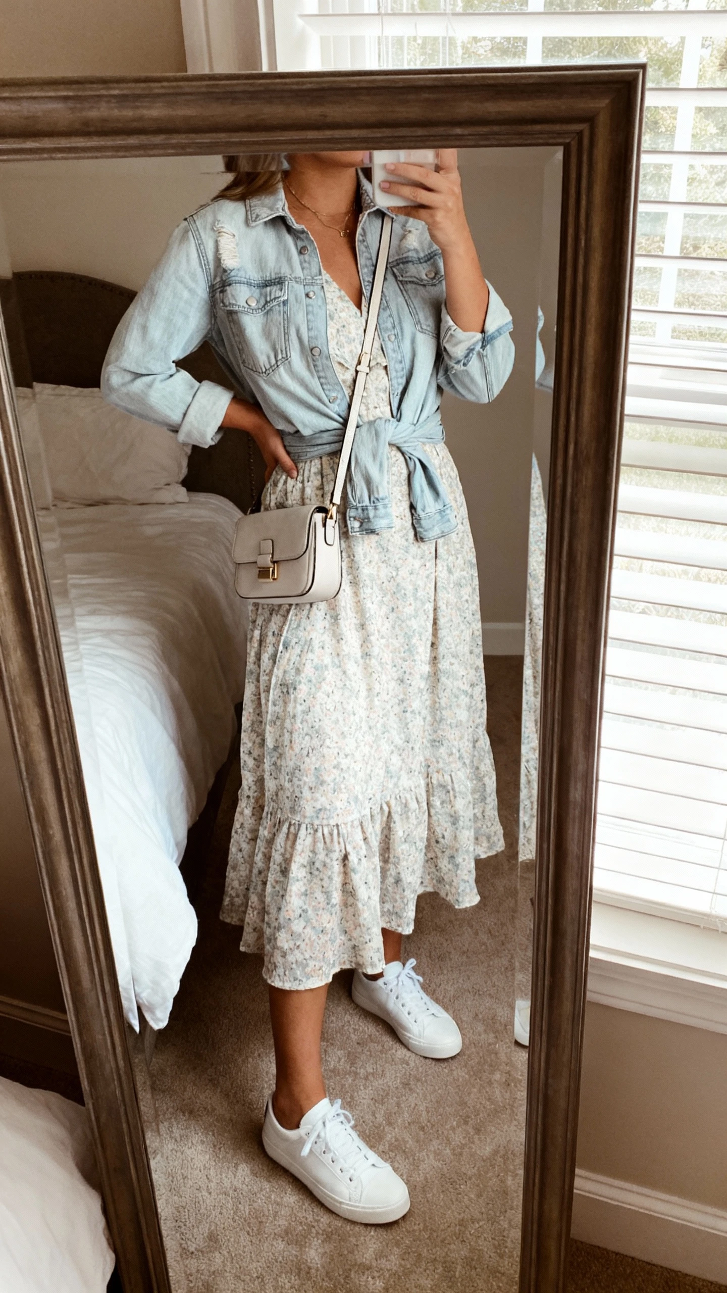 iPhone mirror selfie of a woman wearing a breezy spring dress with white low-top sneakers, a light denim jacket tied at the waist, and a small crossbody bag, face not visible, cropped at neck, bedroom mirror with natural window light, iPhone photo quality.