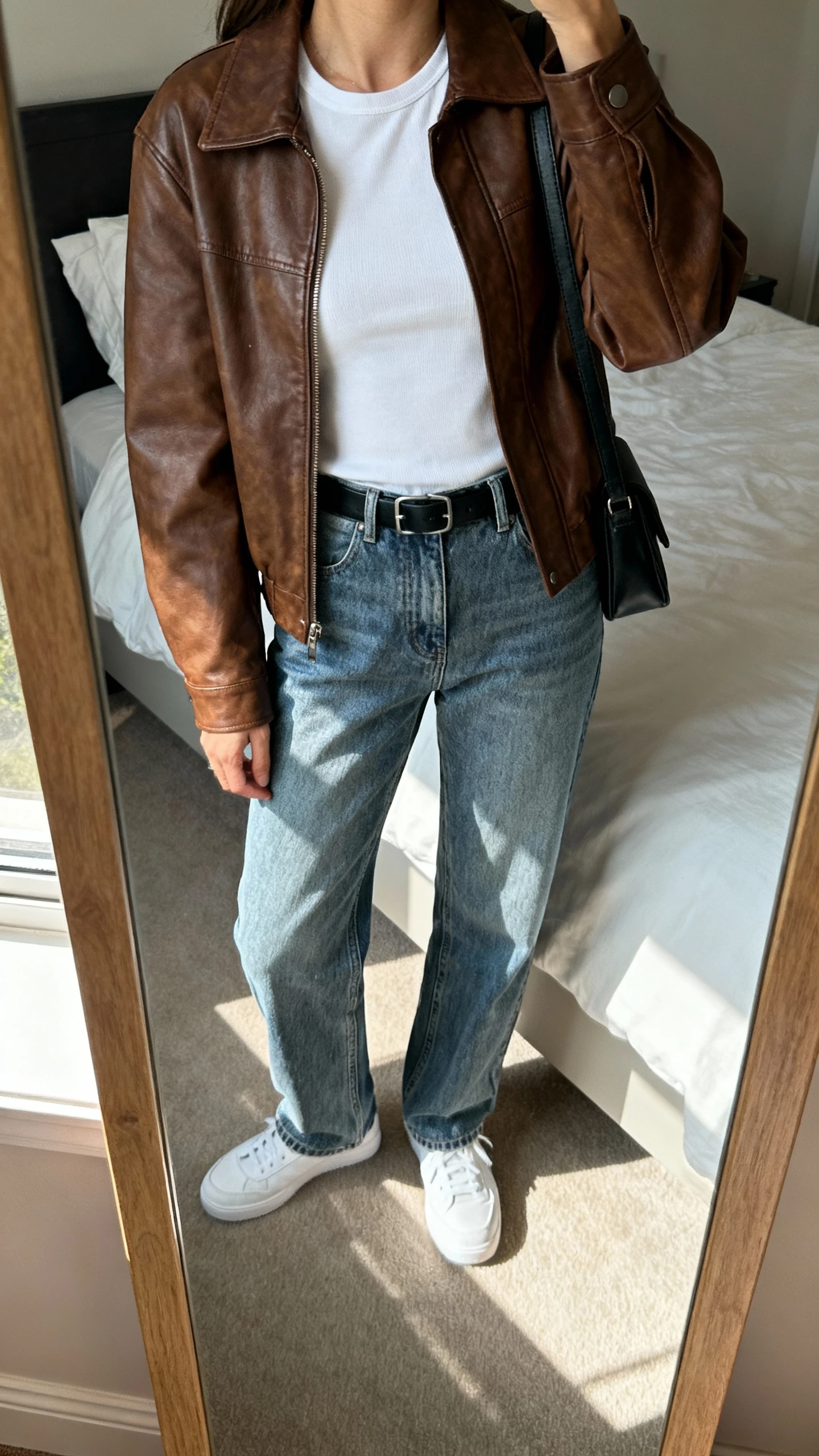iPhone mirror selfie of a woman wearing a brown leather jacket over a white fitted tee, straight-leg medium-wash crisp denim jeans, minimal black belt, white sneakers, and a simple black crossbody, face not visible, cropped at neck, bedroom mirror with natural window light, iPhone photo quality.