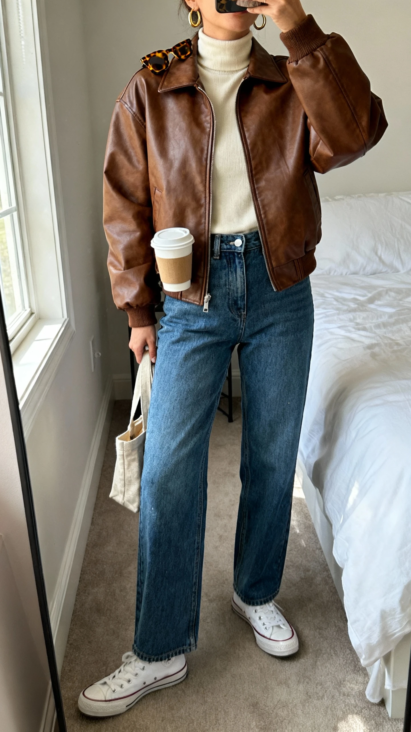iPhone mirror selfie of a woman wearing a brown leather bomber jacket over a cream turtleneck, high-waisted straight-leg blue jeans, white retro sneakers, thin gold hoops, tortoiseshell sunglasses perched on head, and carrying a small canvas tote with a takeaway coffee, face not visible, cropped at neck, bedroom mirror, soft morning window light, iPhone photo quality
