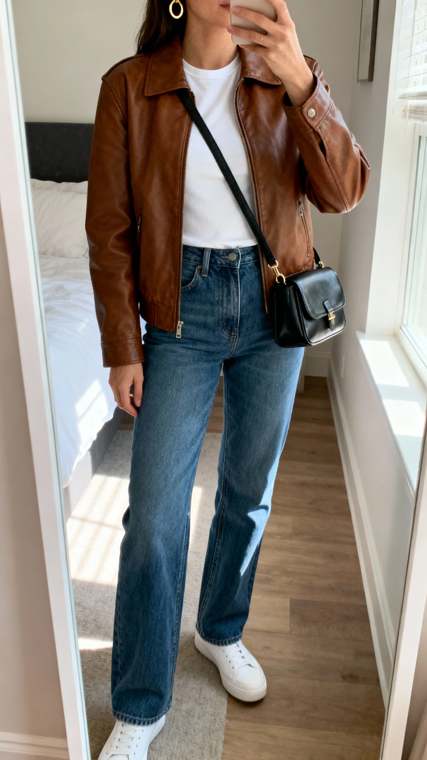 iPhone mirror selfie of a woman wearing a brown leather jacket over a white fitted tee, straight-leg blue jeans, minimalist white sneakers, and a small black leather crossbody with simple gold hoops, face not visible, cropped at neck, bedroom mirror, morning window light, iPhone photo quality.