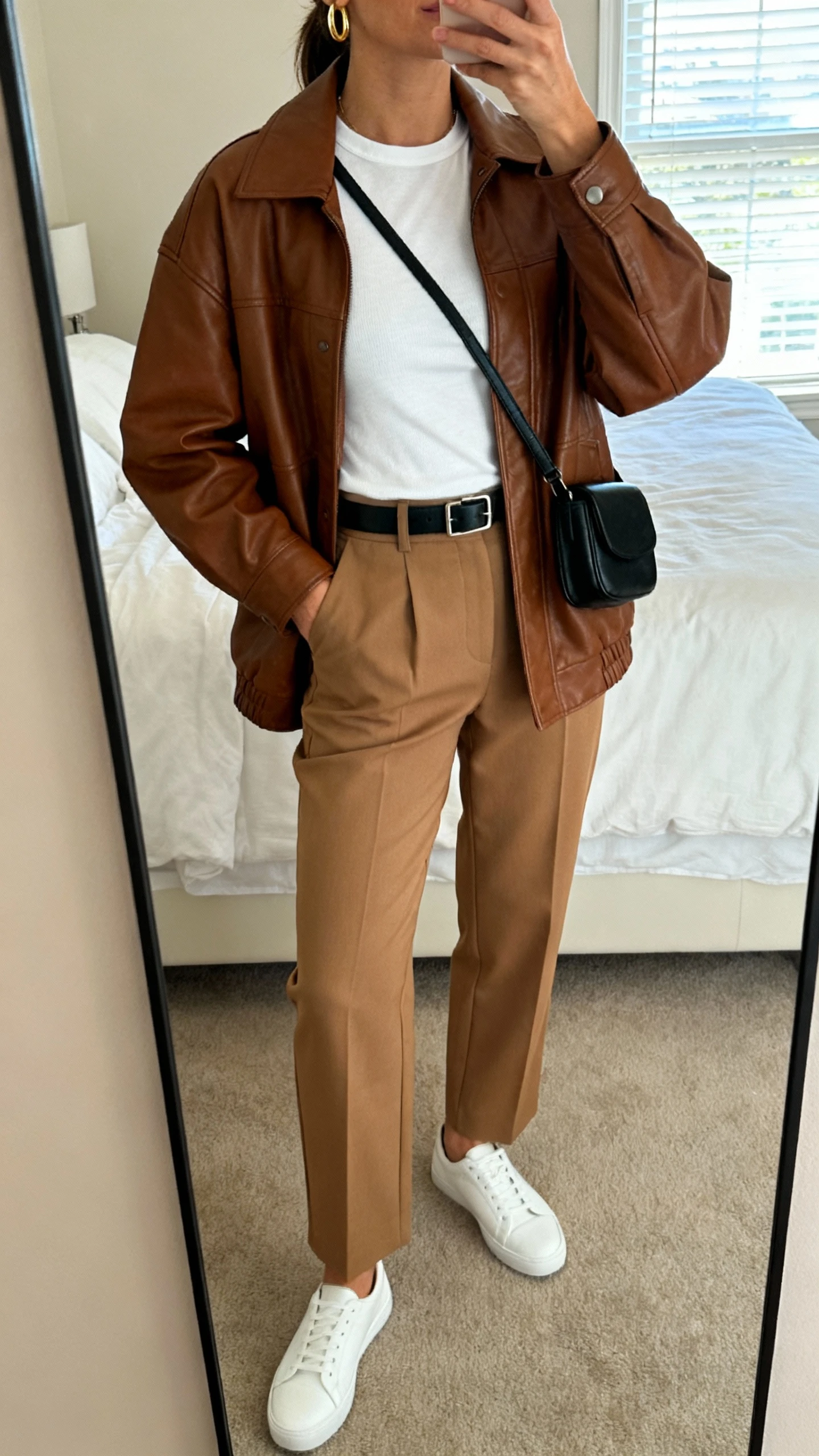 iPhone mirror selfie of a woman wearing a brown leather jacket over a white fitted tee, high-waisted tan tailored trousers, white minimalist sneakers, slim black belt, small black crossbody, simple gold hoops, face not visible, cropped at neck, bedroom mirror, soft morning window light, iPhone photo quality