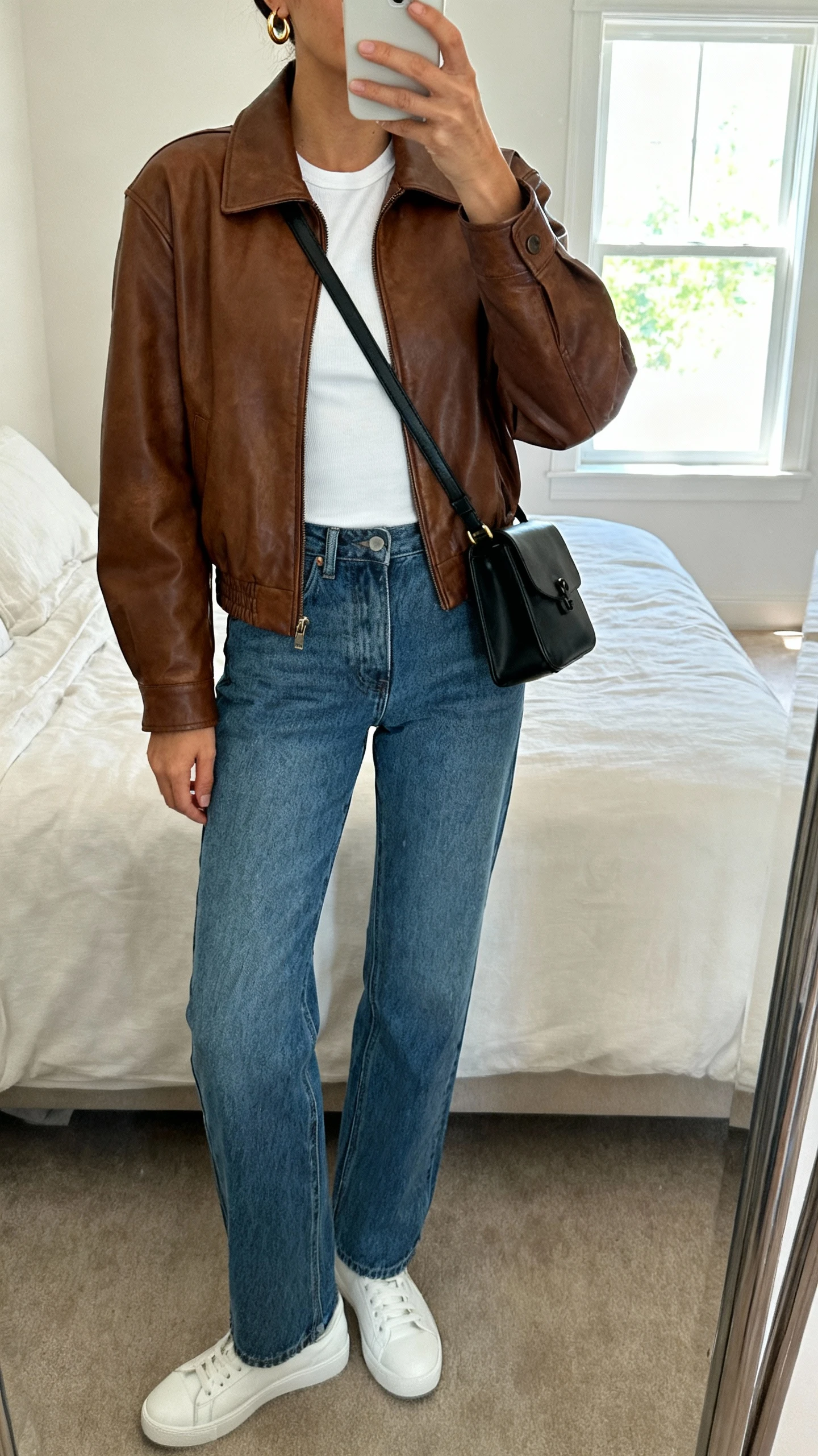 iPhone mirror selfie of a woman wearing a brown leather jacket over a white fitted tee, straight-leg blue jeans, white sneakers, a black crossbody bag, and minimal gold hoop earrings, face not visible, cropped at neck, bedroom mirror with natural window light, iPhone photo quality.
