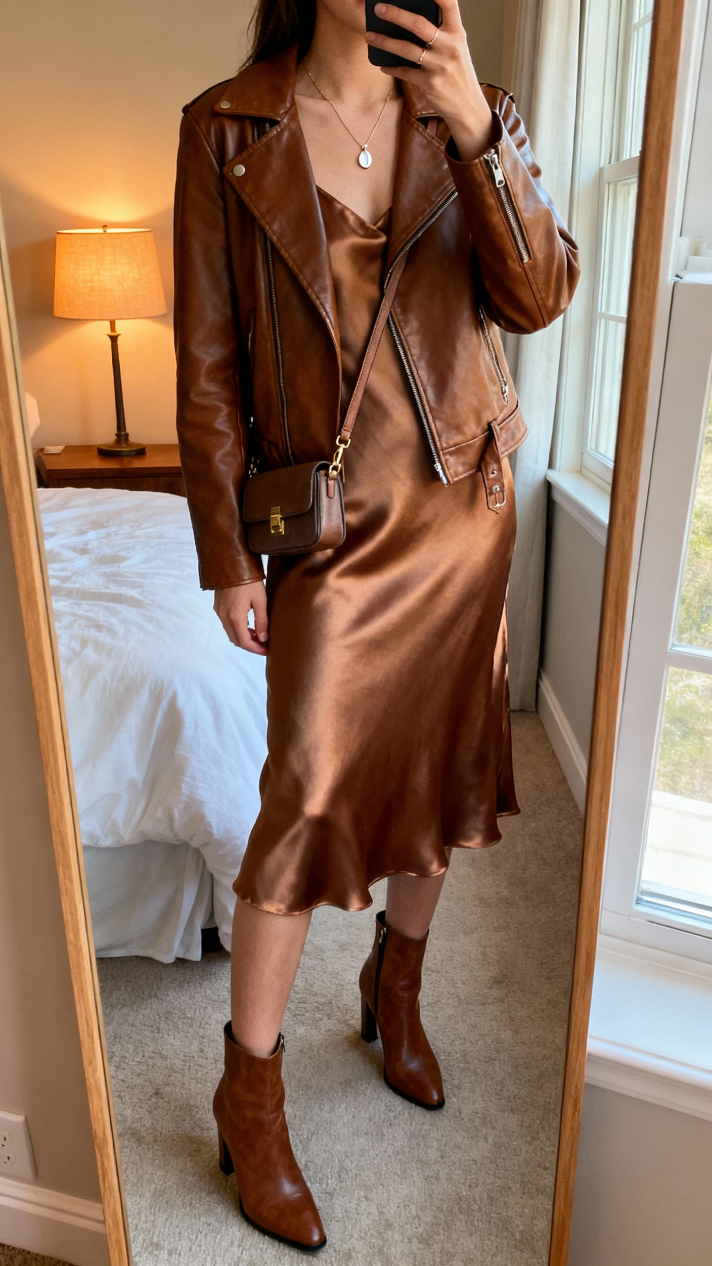 iPhone mirror selfie of a woman wearing a brown leather jacket layered over a satin slip dress with heeled ankle boots, delicate pendant necklace, small crossbody bag, face not visible, cropped at neck, bedroom mirror, warm lamp and window light, iPhone photo quality