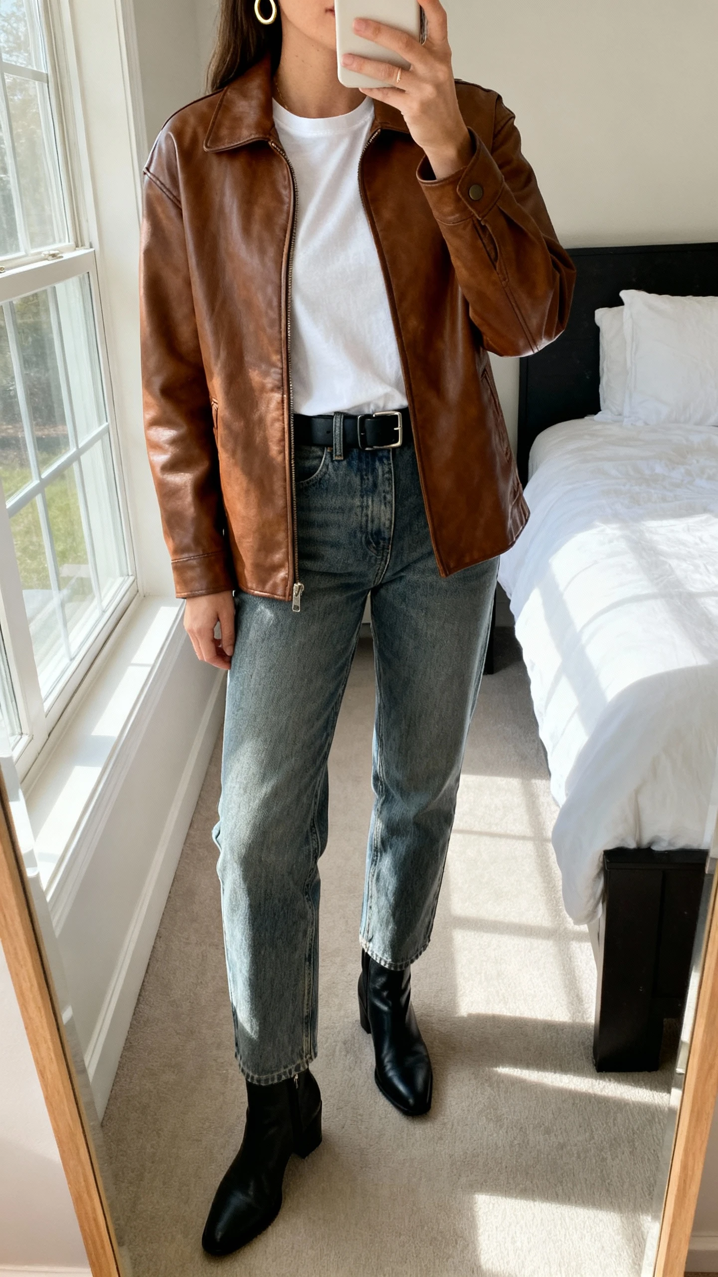 iPhone mirror selfie of a woman wearing a brown leather jacket over a white tee with straight-leg medium-wash denim, black ankle boots, simple black belt, minimal gold hoops, face not visible, cropped at neck, bedroom mirror, natural window light, iPhone photo quality