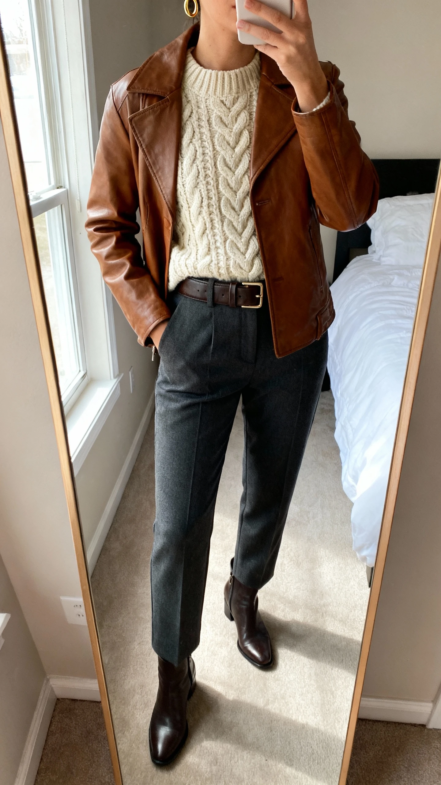 iPhone mirror selfie of a woman wearing a brown leather jacket over a chunky cream cable-knit sweater, tailored charcoal trousers, sleek ankle boots, a structured leather belt, and minimal gold hoops, face not visible, cropped at neck, bedroom mirror with natural window light, iPhone photo quality.