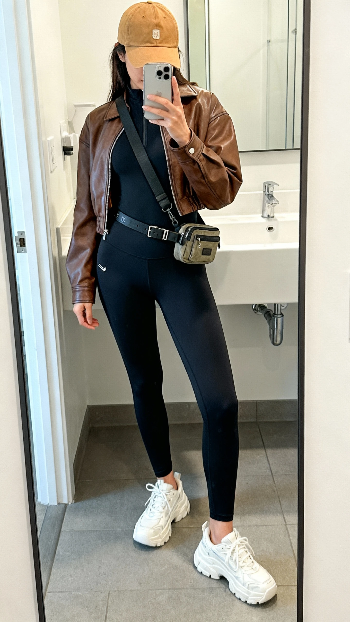 iPhone mirror selfie of a woman wearing a brown leather jacket dupe over a longline black athletic top, high-waisted black leggings, chunky white sneakers, baseball cap in camel, and a belt bag worn crossbody, face not visible, cropped at shoulders, bathroom mirror, bright indoor lighting, iPhone photo quality