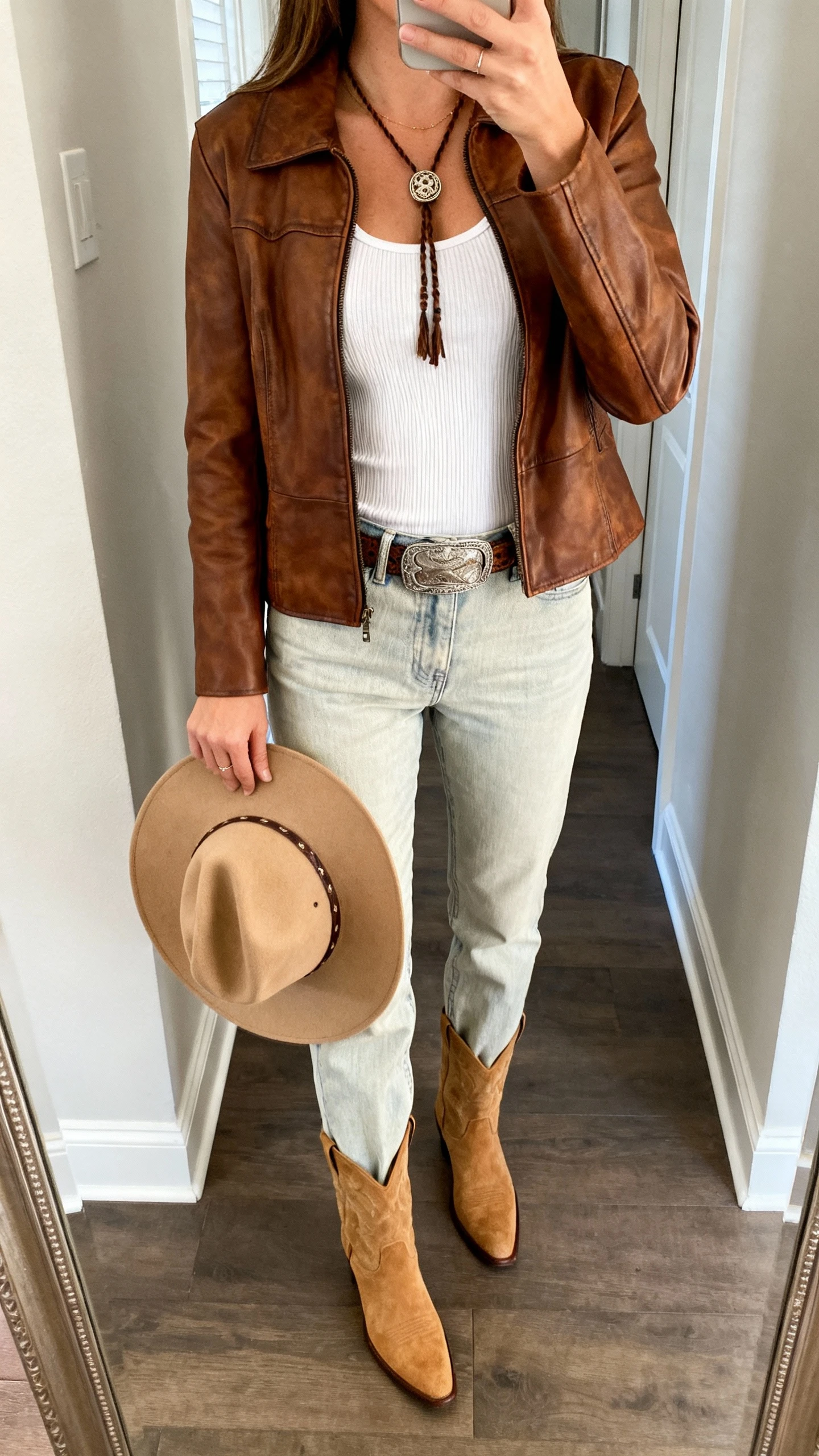 iPhone mirror selfie of a woman wearing a brown leather jacket with a white ribbed tank, light-wash straight-leg jeans, western belt, tan suede ankle boots, bolo-style necklace, camel felt cowboy hat in hand, face not visible, cropped at neck, hallway full-length mirror, natural daylight, iPhone photo quality