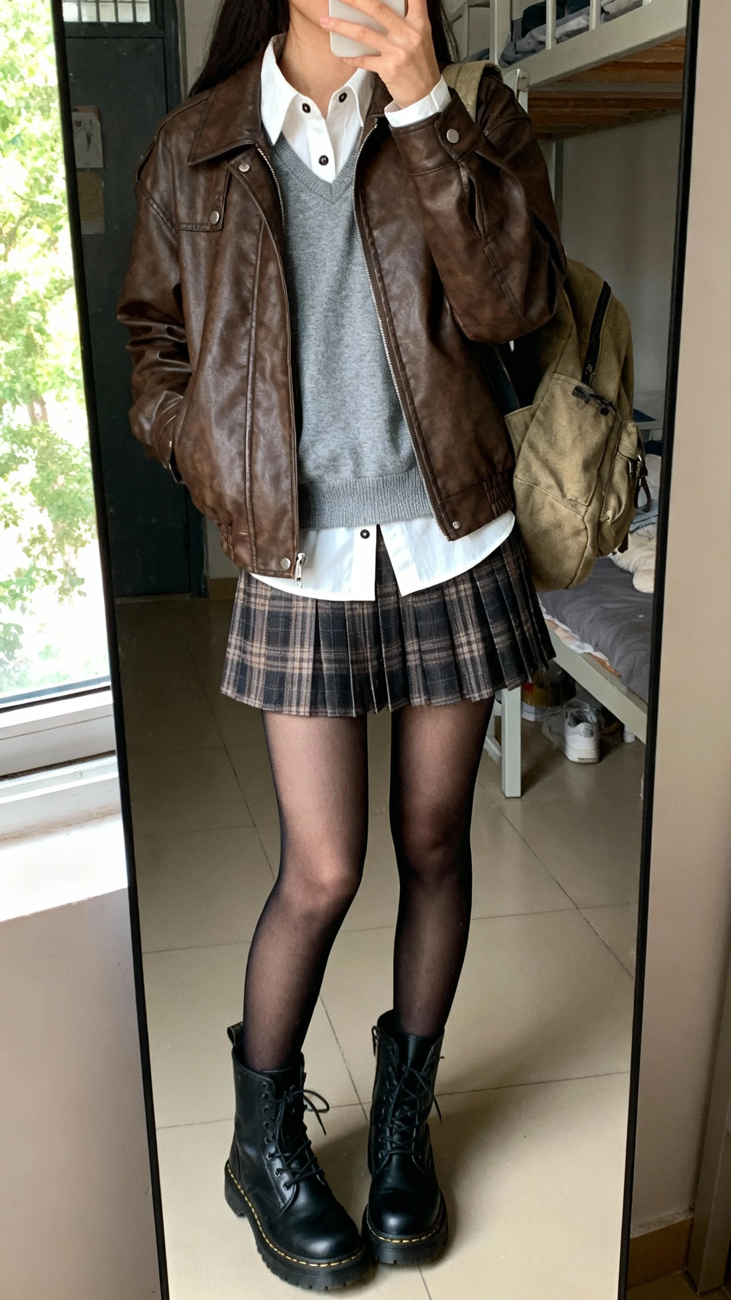 iPhone mirror selfie of a woman wearing a brown leather moto jacket over a grey crewneck sweater layered atop a white button-up shirt with the collar peeking, pleated plaid mini skirt, black sheer tights, black combat boots, and a canvas backpack, face not visible, cropped at shoulders, dorm bedroom mirror, afternoon window light, iPhone photo quality.