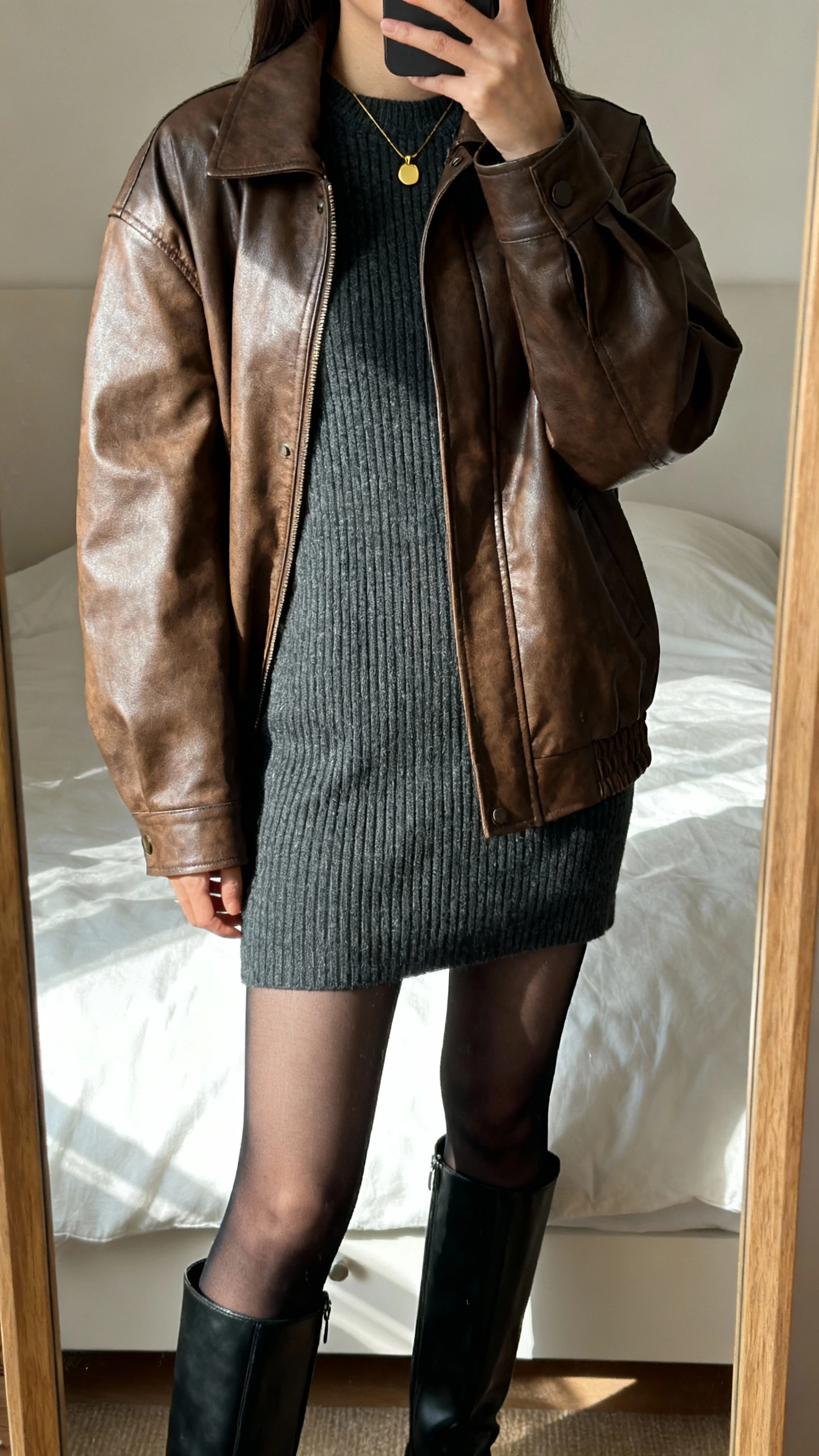iPhone mirror selfie of a woman wearing a brown leather jacket layered over a ribbed knit sweater dress in charcoal, sheer black tights, black knee-high boots, and a minimal gold pendant, face not visible, cropped at shoulders, bedroom mirror with soft afternoon light, iPhone photo quality.