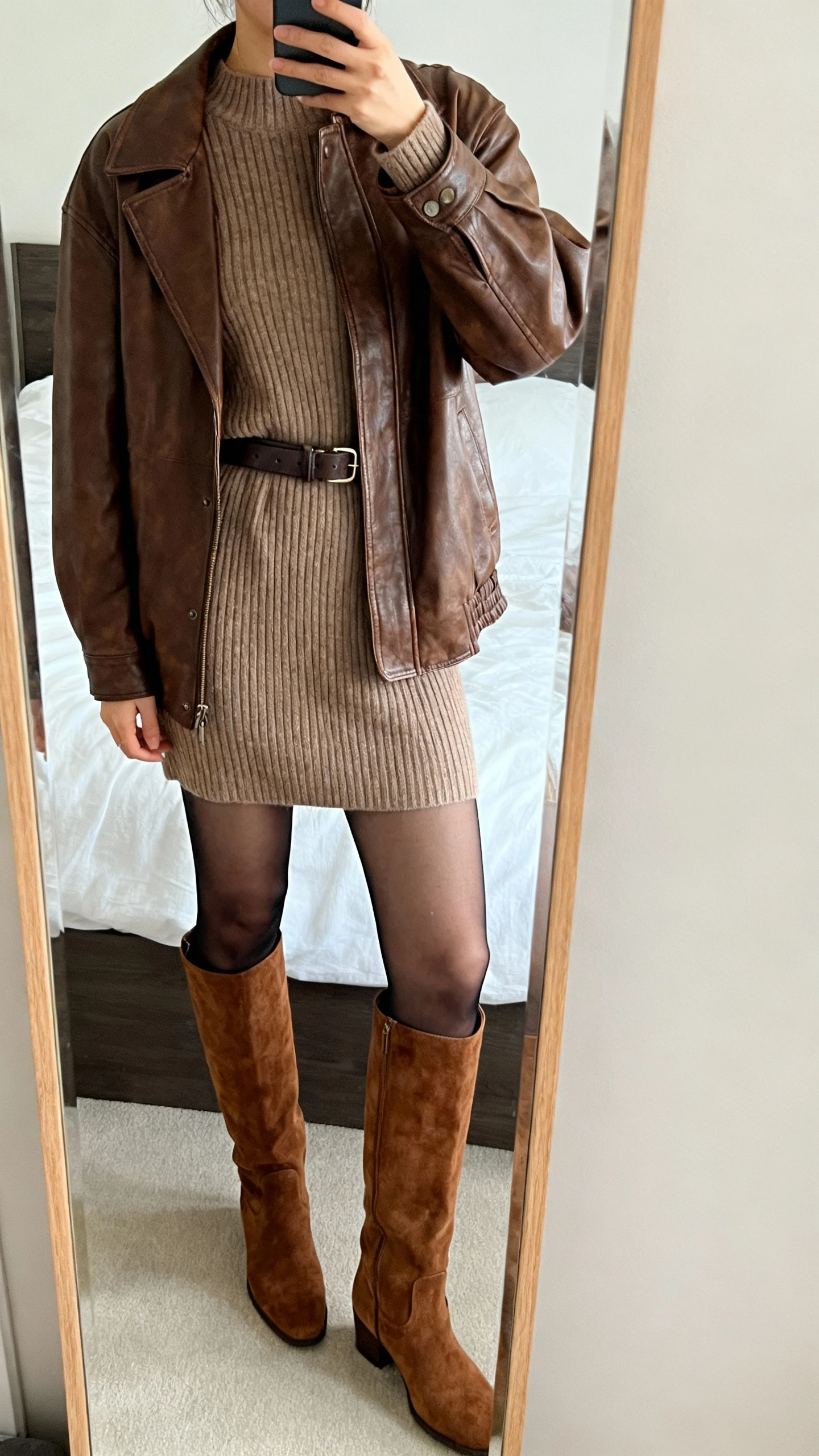 iPhone mirror selfie of a woman wearing a brown leather jacket over a ribbed sweater dress, over-the-knee suede boots, sheer black tights, and a waist belt, face not visible, cropped at shoulders, bedroom mirror, soft natural light, iPhone photo quality.