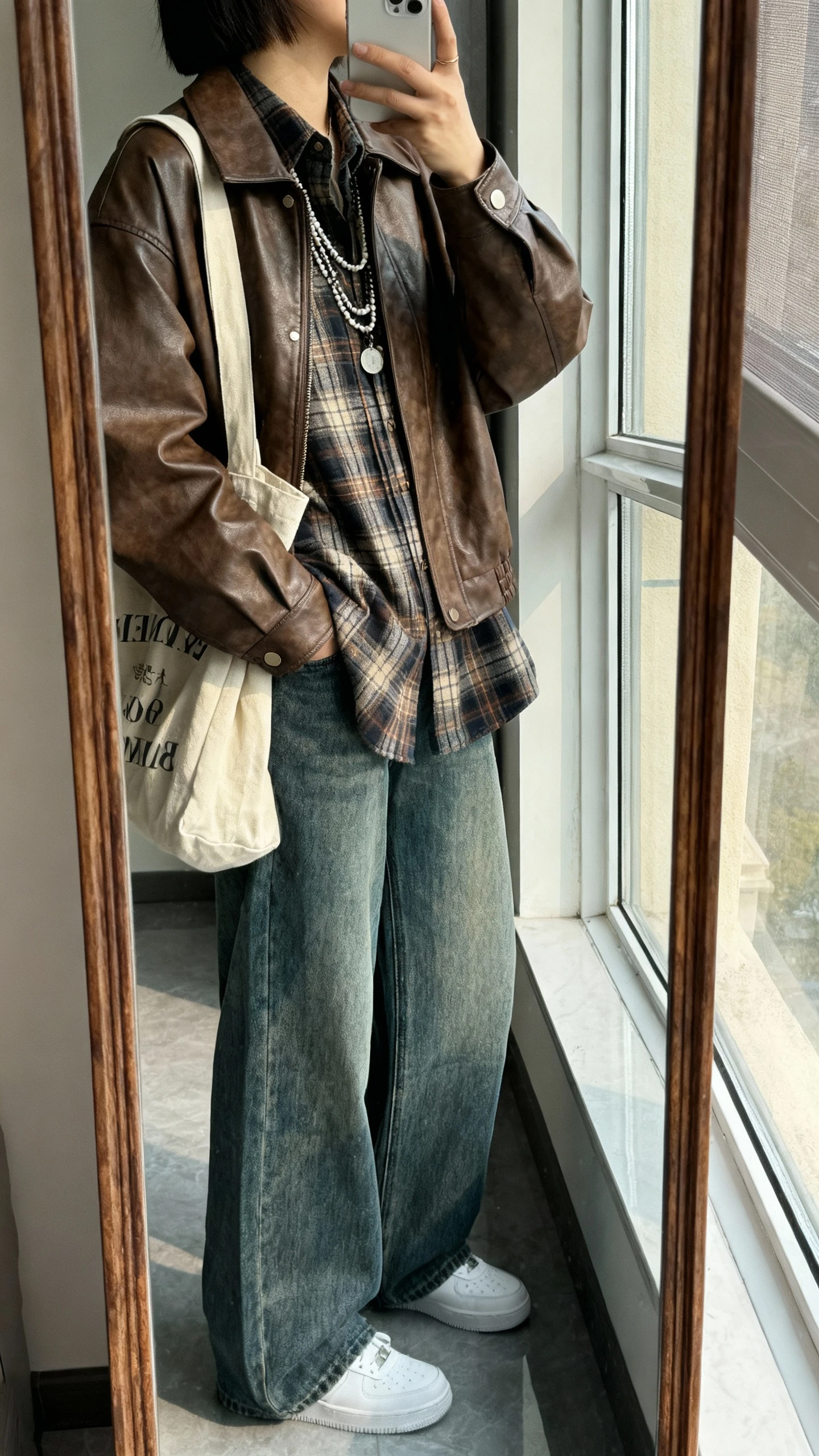 iPhone mirror selfie of a woman wearing a brown leather jacket over an elevated plaid flannel shirt, wide-leg denim, white sneakers, and a canvas tote with layered necklaces, face not visible, side angle, full-length mirror by a window, iPhone photo quality.