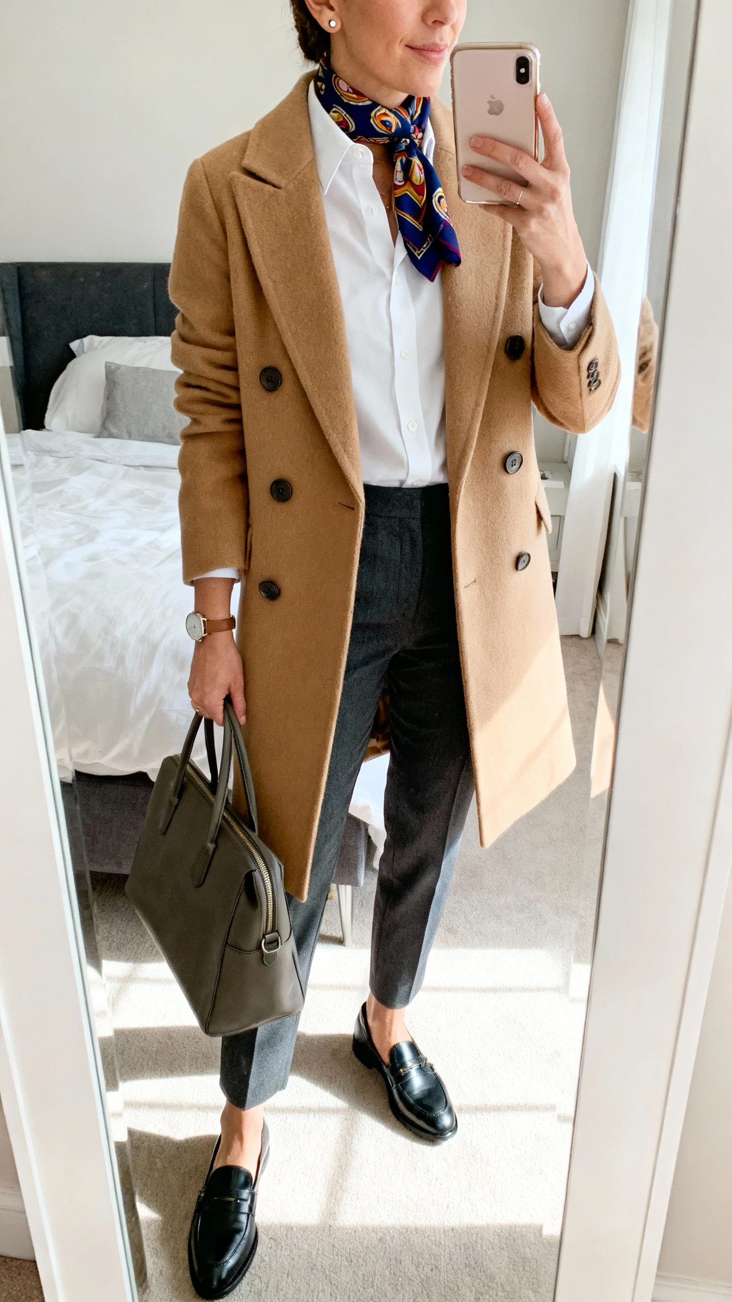 iPhone mirror selfie of a woman wearing a camel double-breasted wool coat over tailored charcoal trousers, a crisp white button-up, polished black leather loafers, a structured work tote, a minimal watch with small stud earrings, and a jewel-toned graphic scarf, face not visible, cropped at neck, bedroom mirror, natural daylight, iPhone photo quality.
