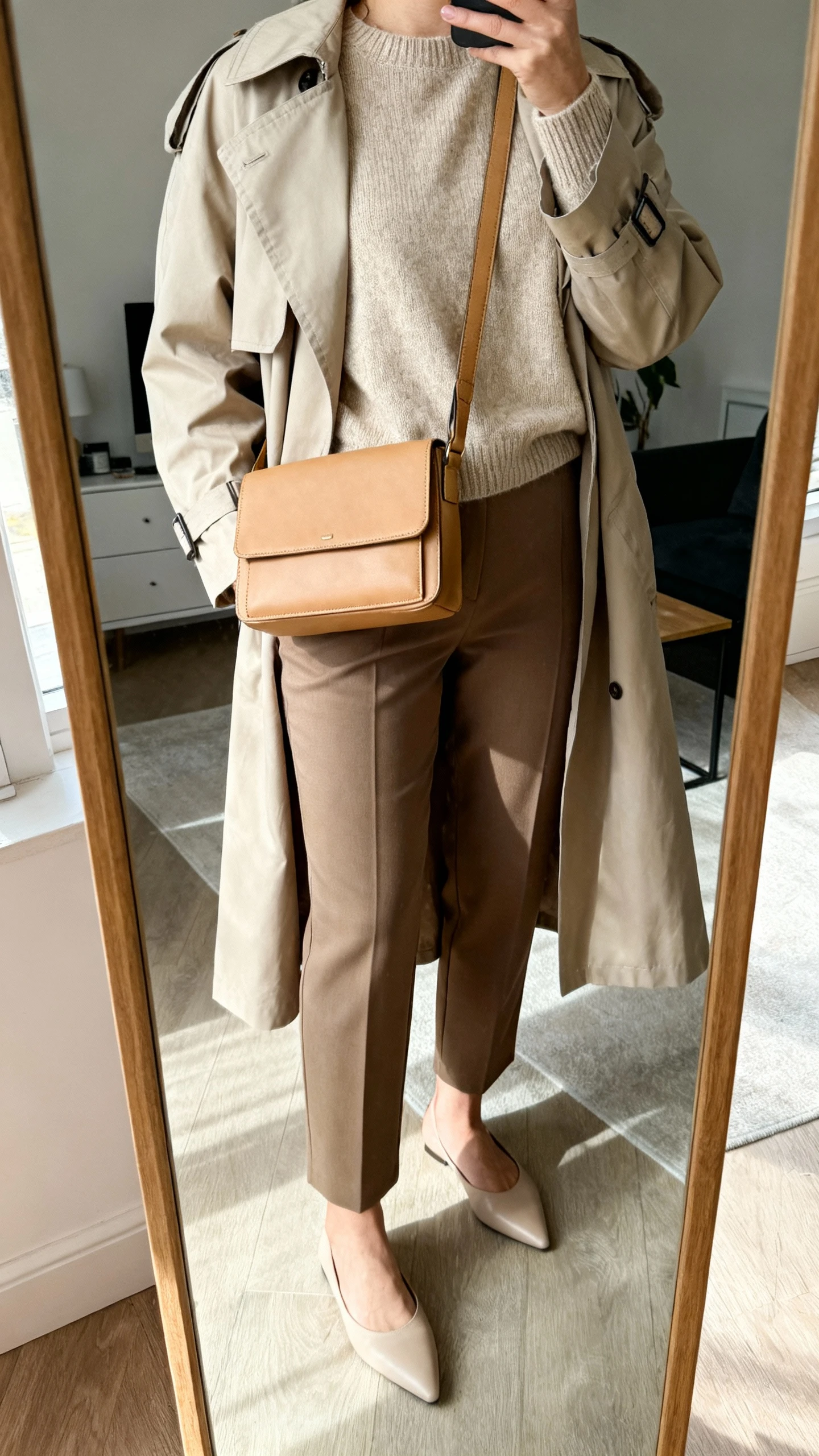 iPhone mirror selfie of a woman wearing a camel structured crossbody with flap closure, a soft merino crewneck sweater, tailored ankle-length trousers, minimal pointed flats, and a lightweight trench coat, face not visible, cropped at shoulders, full-length mirror, indoor natural light, iPhone photo quality.