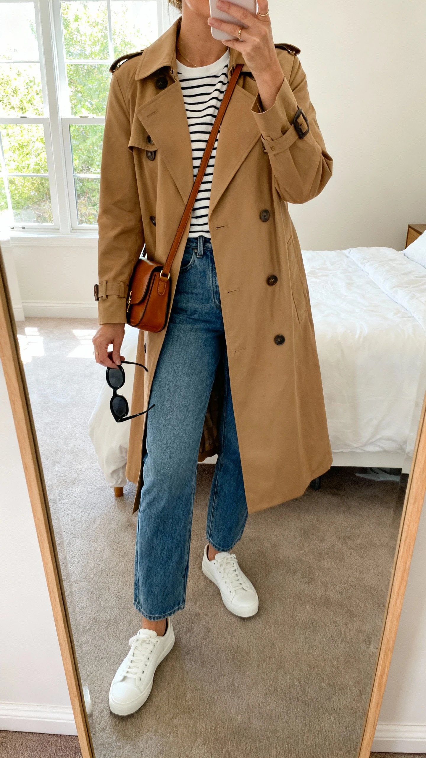 iPhone mirror selfie of a woman wearing a camel trench coat over a striped tee, straight-leg blue jeans, white low-top sneakers, crossbody leather bag, and black sunglasses in hand, face not visible, cropped at neck, bedroom mirror with natural window light, iPhone photo quality.