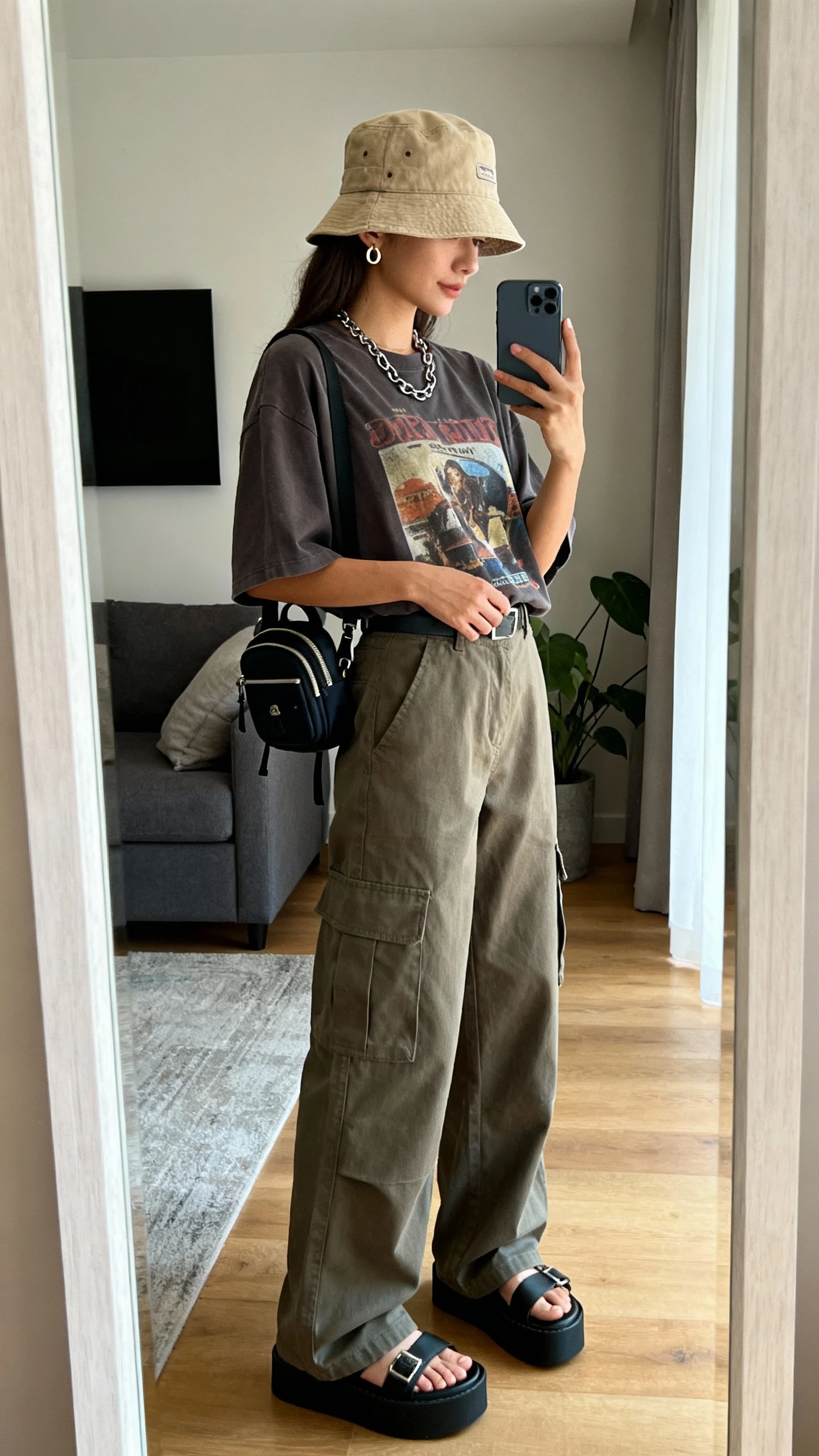 iPhone mirror selfie of a woman wearing a canvas bucket hat, boxy graphic tee tucked into wide-leg cargo pants, platform sandals, layered chain necklace, tiny hoop earrings, mini backpack, face not visible, side angle, living room full-length mirror, natural daylight, iPhone photo quality.