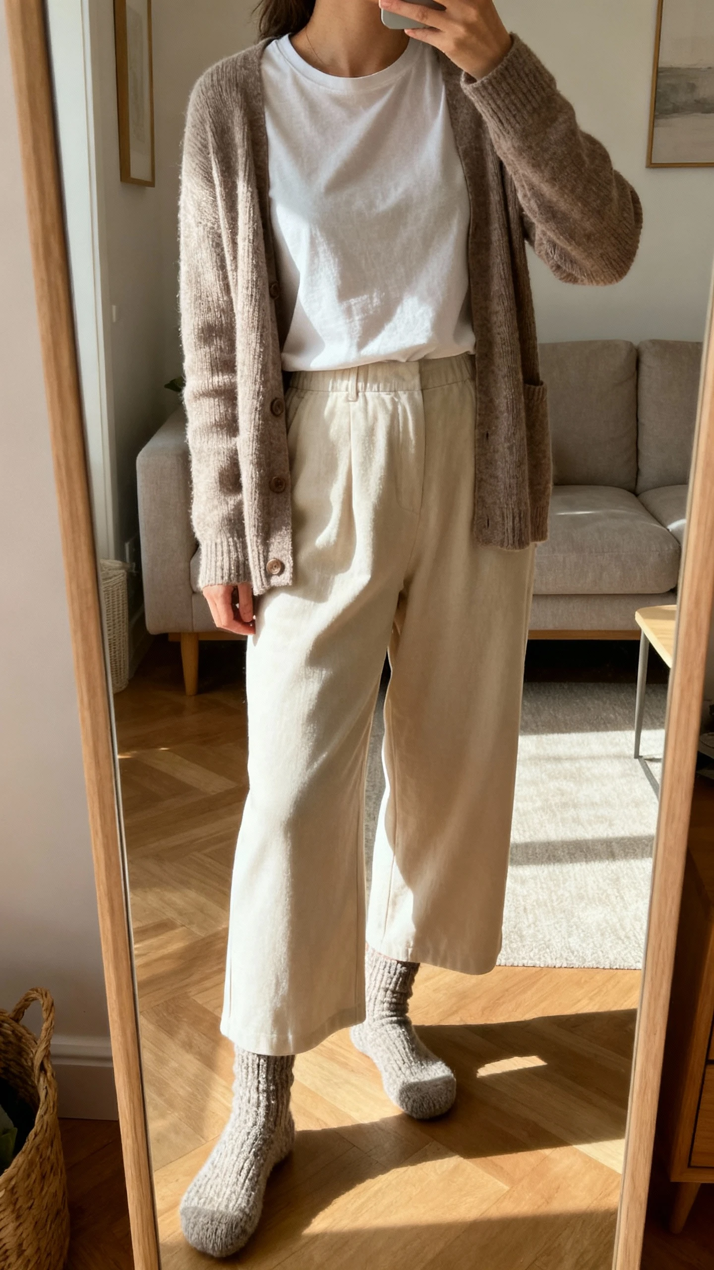 iPhone mirror selfie of a woman wearing a cardigan layered over a plain tee with soft wide-leg pants and cozy socks, face not visible, cropped at neck, living room mirror, gentle morning light, iPhone photo quality.
