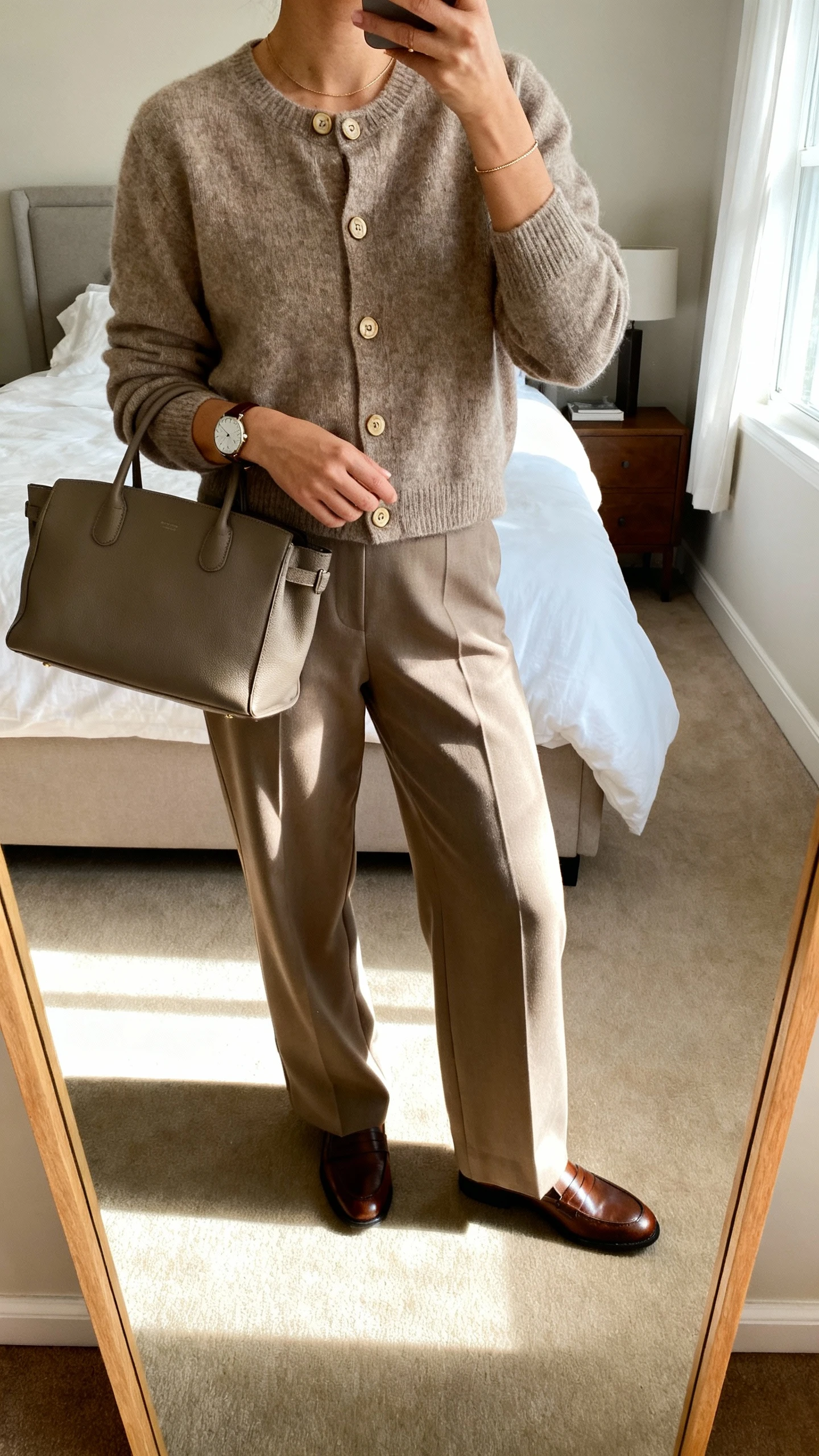 iPhone mirror selfie of a woman wearing a cashmere cardigan (buttoned), wide-leg trousers, and leather loafers, with a structured tote and delicate watch, face not visible, cropped at neck, bedroom mirror, soft afternoon light, iPhone photo quality.