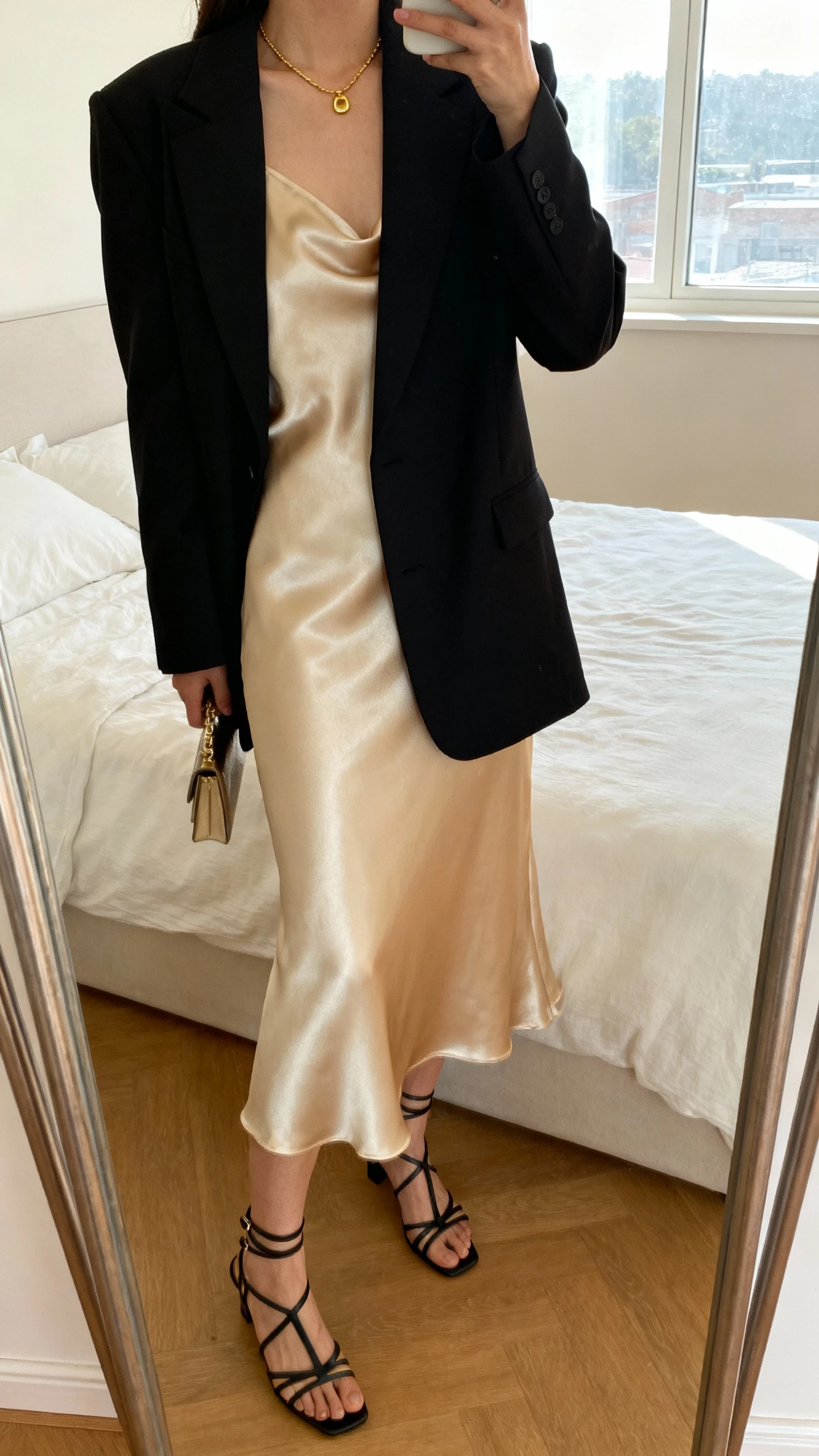 iPhone mirror selfie of a woman wearing a champagne satin slip dress layered with a tailored black blazer, delicate gold necklace, strappy heels, and a small clutch, face not visible, cropped at neck, bedroom mirror with natural window light, iPhone photo quality.