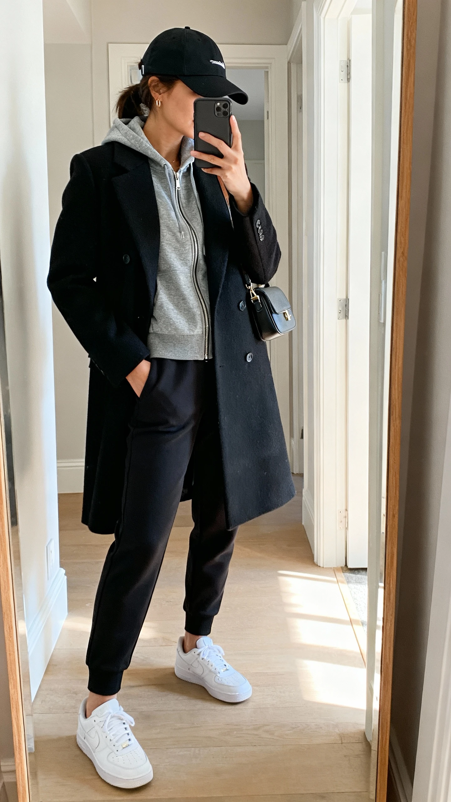 iPhone mirror selfie of a woman wearing a chic athleisure mix: black tailored wool coat over a grey zip-up hoodie, black tapered joggers, clean white sneakers, and a sporty black cap with a small shoulder bag, face not visible, side angle, entryway mirror, natural daylight, iPhone photo quality.