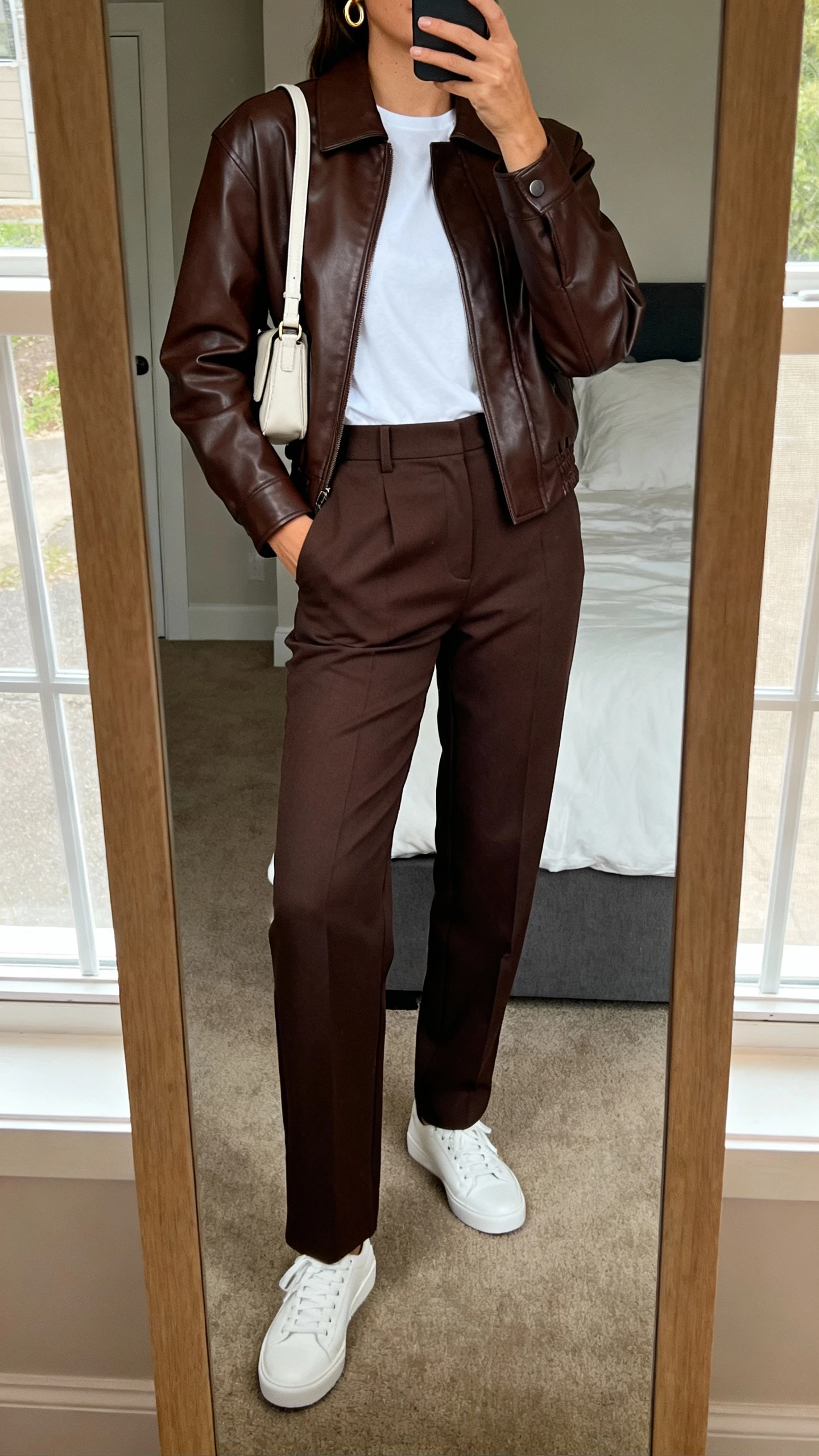 iPhone mirror selfie of a woman wearing a chocolate leather jacket, tailored high-waist trousers, sleek white low-top sneakers, simple white tee, minimalist crossbody bag, and subtle gold hoops, face not visible, cropped at neck, bedroom mirror with natural window light, iPhone photo quality.
