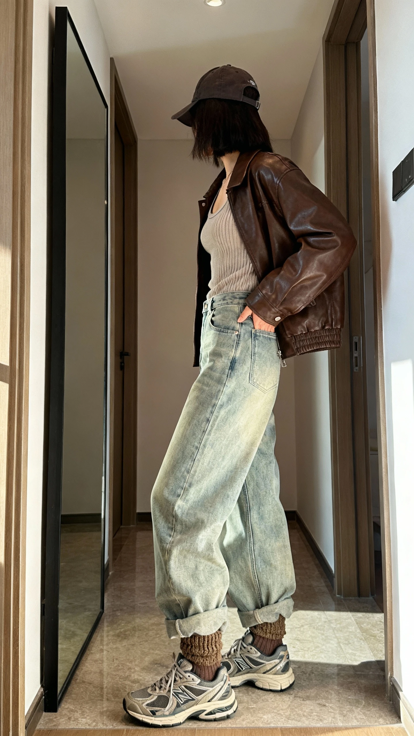 iPhone mirror selfie of a woman wearing a chocolate leather jacket, baggy light-wash denim jeans, retro trainers, ribbed tank, baseball cap, and chunky socks peeking, face not visible, side angle cropped at shoulders, hallway full-length mirror, morning daylight, iPhone photo quality.