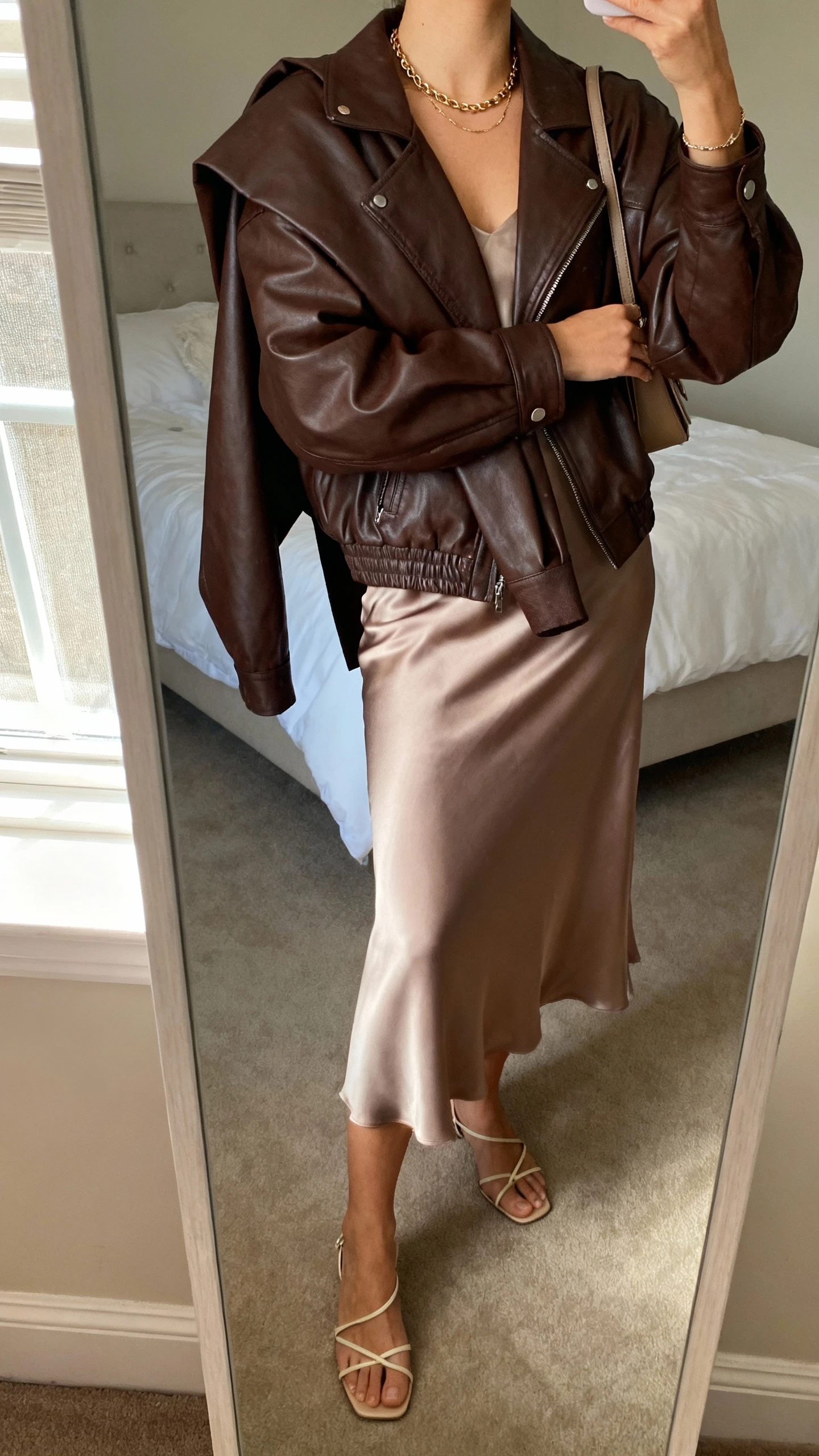 iPhone mirror selfie of a woman wearing a chocolate leather jacket draped over a silk midi skirt, minimal strappy heels, fine chain jewelry, and a slim shoulder bag, face not visible, cropped at neck, bedroom mirror with soft window light, iPhone photo quality.