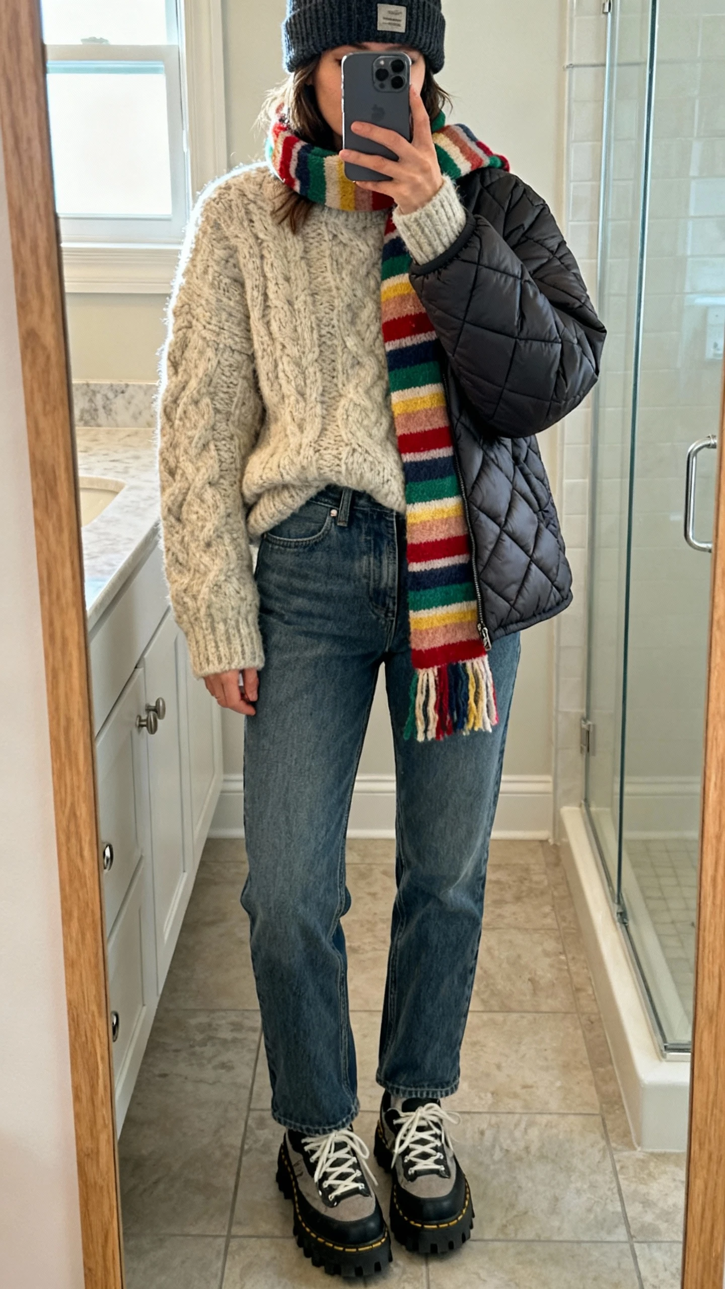 iPhone mirror selfie of a woman wearing a chunky cozy knit sweater, relaxed-fit jeans, a quilted puffer jacket, colorful striped scarf looped loosely, beanie, and lug-sole sneakers, face not visible, bathroom mirror, soft indoor morning light, iPhone photo quality.