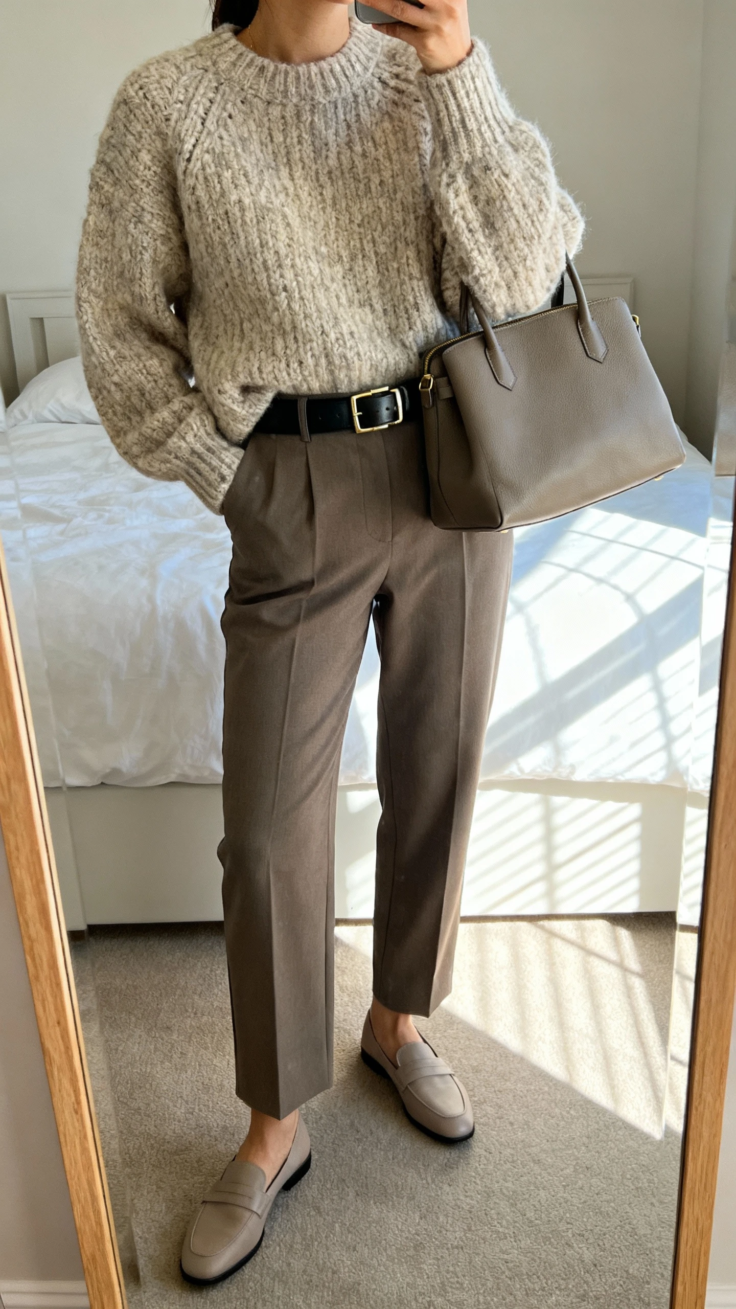 iPhone mirror selfie of a woman wearing a chunky knit sweater, tailored trousers, sleek leather belt, minimalist loafers, and a structured tote, face not visible, cropped at neck, bedroom mirror with natural window light, iPhone photo quality.