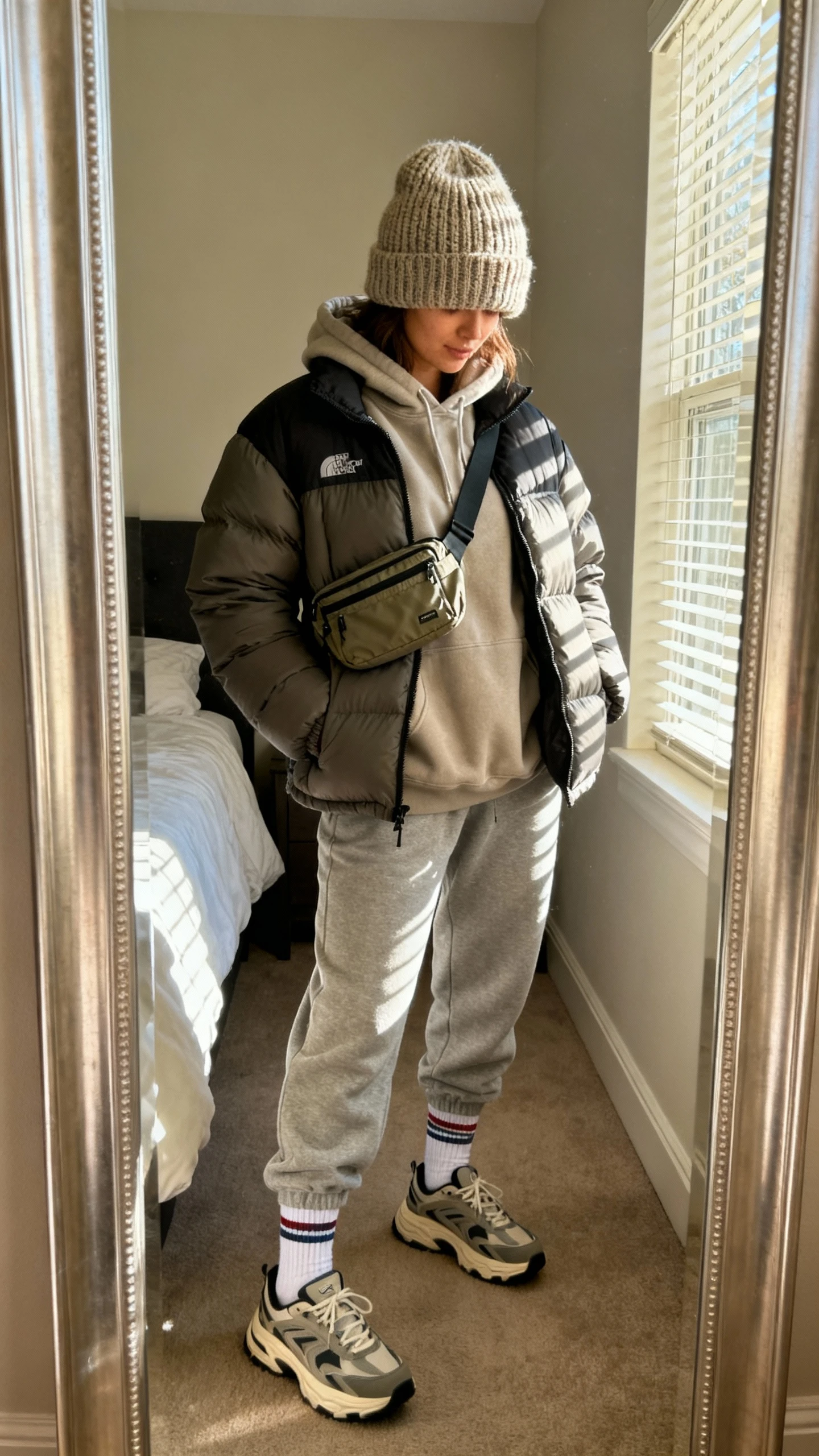 iPhone mirror selfie of a woman wearing a chunky knit beanie, oversized hoodie layered under a puffer jacket, joggers, chunky dad sneakers, crew socks, crossbody nylon belt bag, face not visible, cropped at shoulders, bedroom mirror with afternoon window light, iPhone photo quality.