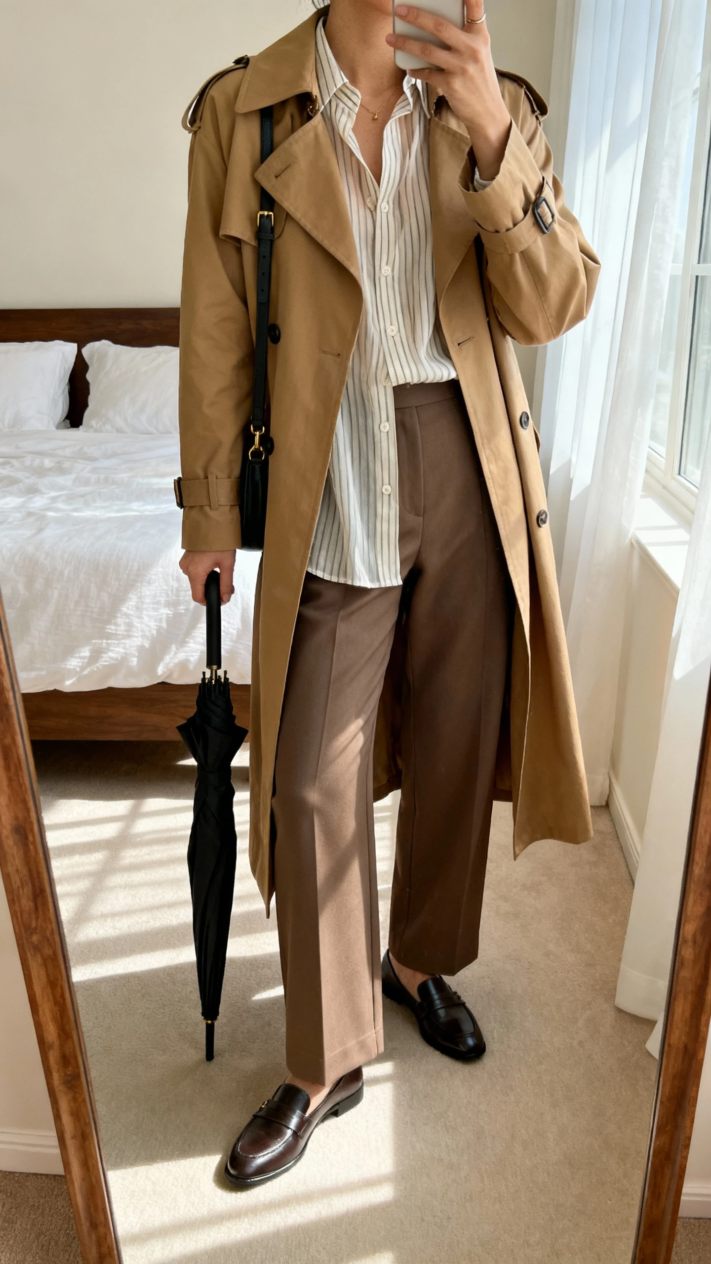 iPhone mirror selfie of a woman wearing a classic tan trench coat over a breezy striped button-up and tailored wide-leg trousers with sleek loafers, black mini umbrella in hand, crossbody bag, face not visible, cropped at neck, bedroom mirror, morning window light, iPhone photo quality.