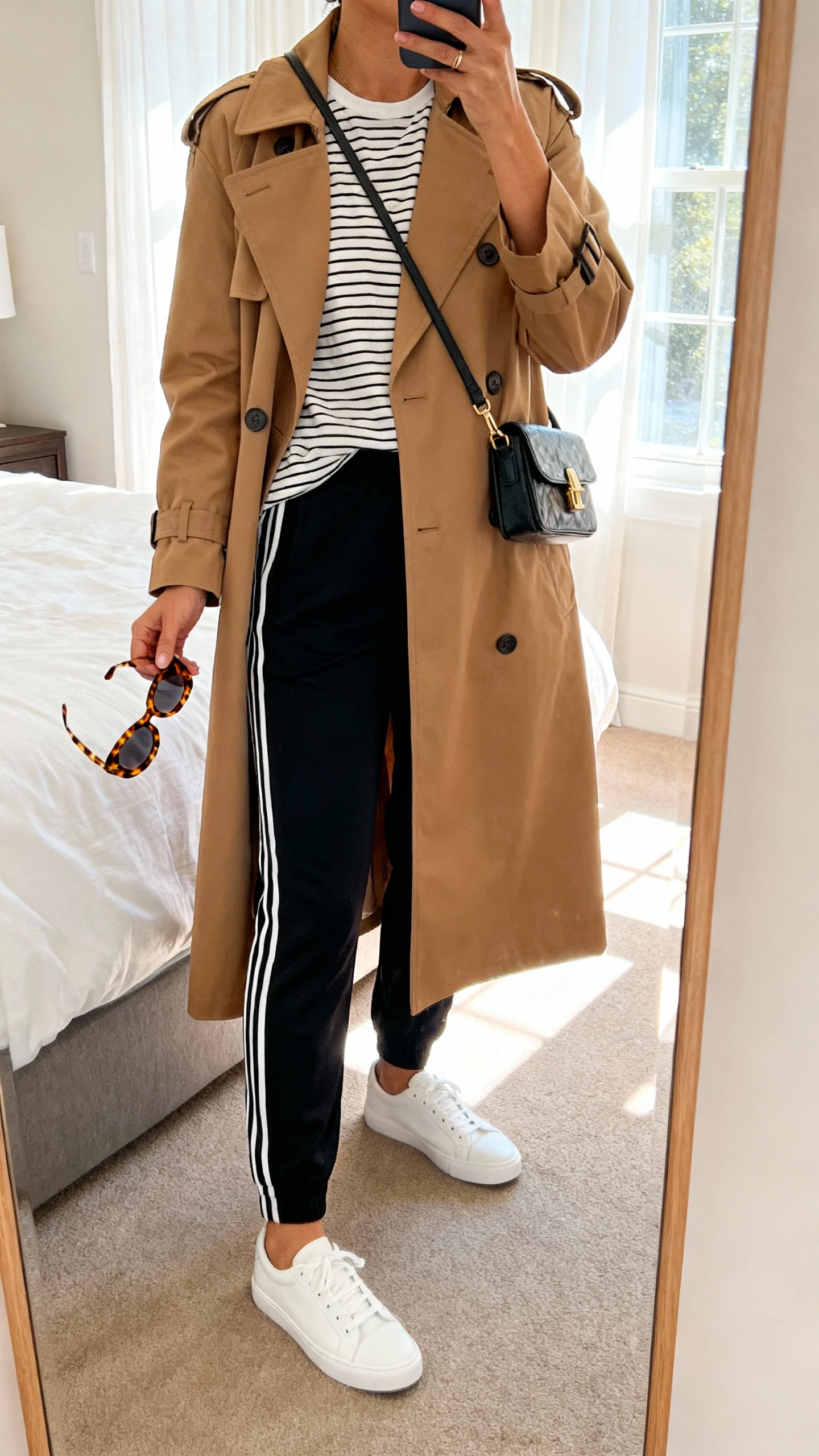 iPhone mirror selfie of a woman wearing a classic camel trench coat over a striped tee, sleek black track pants with side stripes, white low-top sneakers, small crossbody bag, and tortoiseshell sunglasses in hand, face not visible, cropped at neck, bedroom mirror with morning window light, iPhone photo quality.