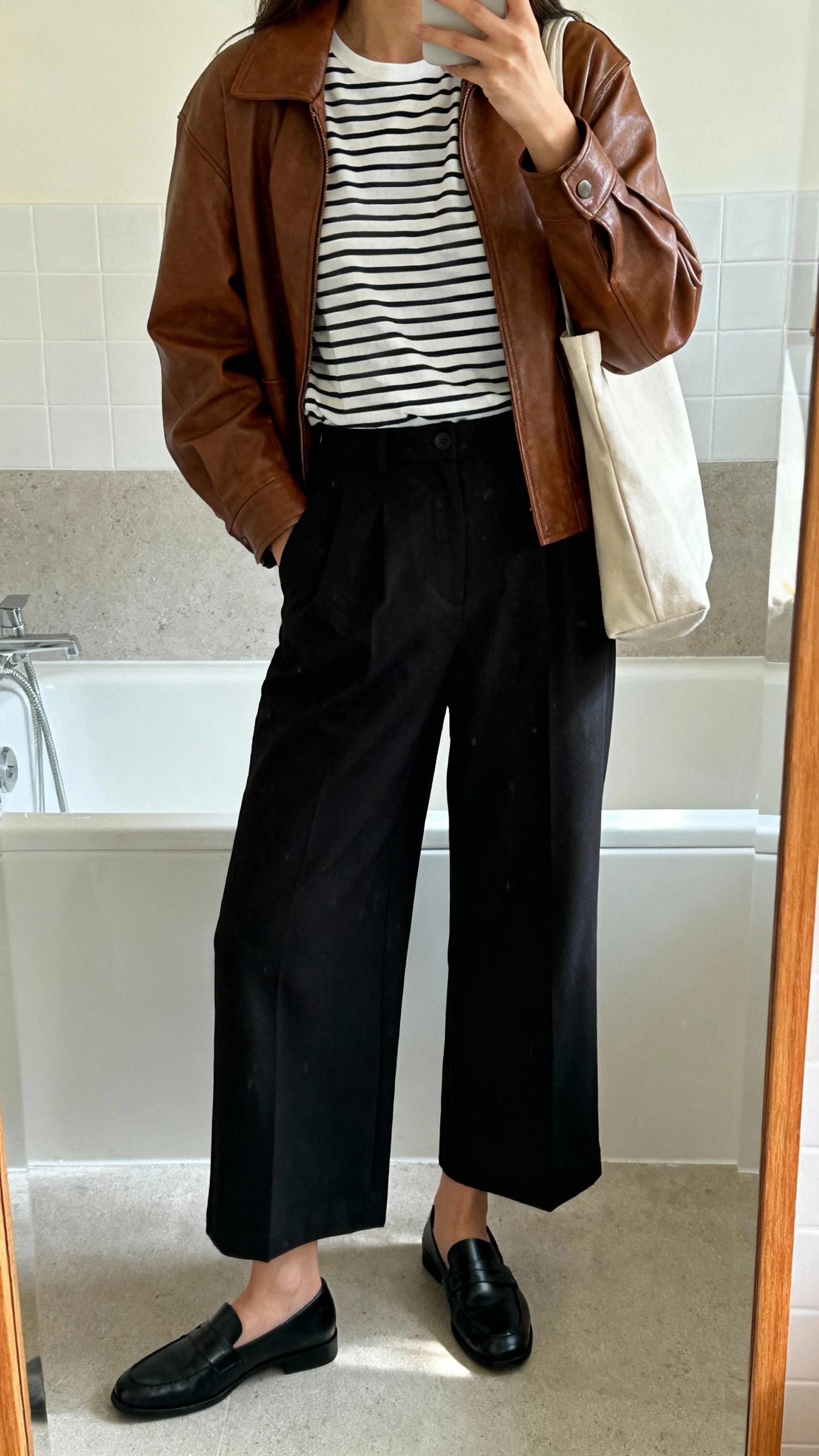 iPhone mirror selfie of a woman wearing a classic Breton striped tee, high-waisted wide-leg black trousers, brown leather jacket, black loafers, and a simple tote, face not visible, bathroom mirror, morning light, iPhone photo quality
