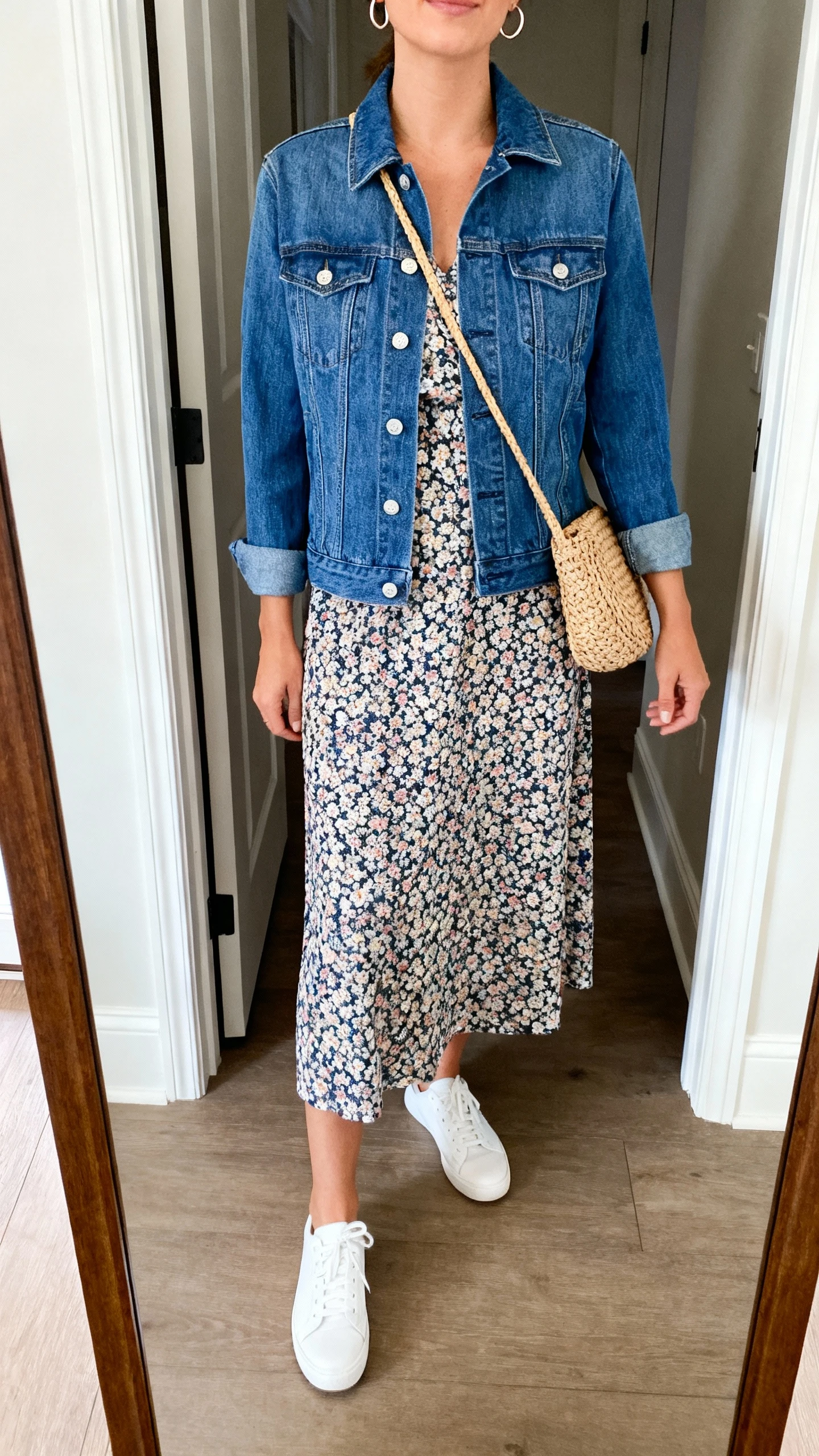 iPhone mirror selfie of a woman wearing a classic blue denim jacket layered over a floral midi dress, white low-top sneakers, small hoop earrings, and a woven crossbody bag, face not visible, cropped at neck, entryway full-length mirror with natural daylight, iPhone photo quality.
