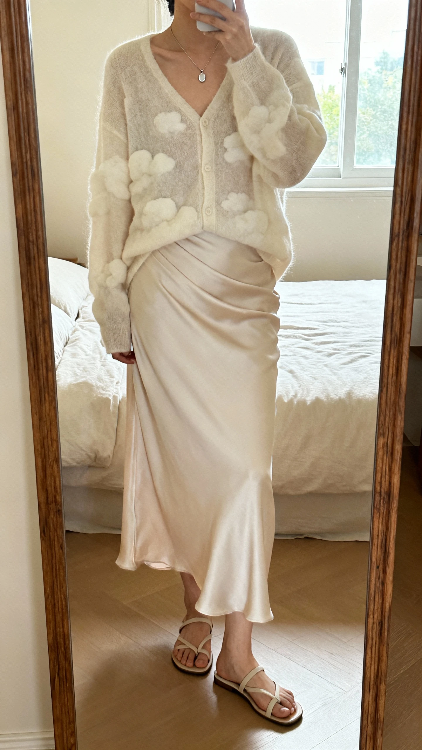 iPhone mirror selfie of a woman wearing a cloud-soft cardigan over a bias-cut slip skirt that moves, delicate pendant necklace, minimalist sandals, face not visible, cropped at neck, bedroom mirror with natural morning window light, iPhone photo quality.