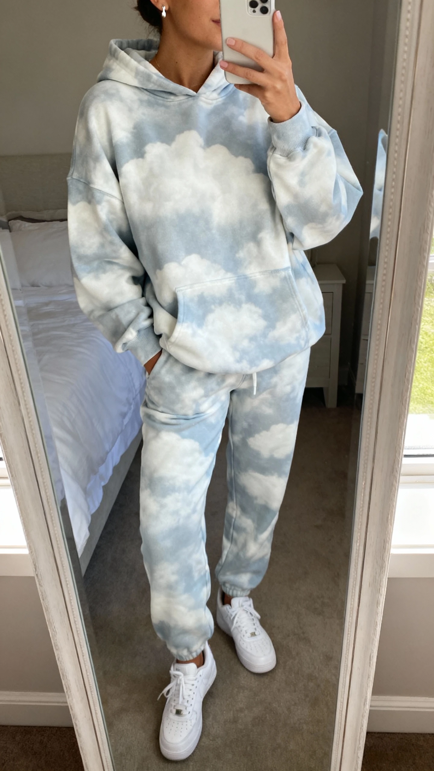 iPhone mirror selfie of a woman wearing a cloud-soft matching sweatsuit set (relaxed hoodie and joggers) with clean white sneakers and simple stud earrings, face not visible, cropped at neck, bedroom mirror, natural window light, iPhone photo quality.