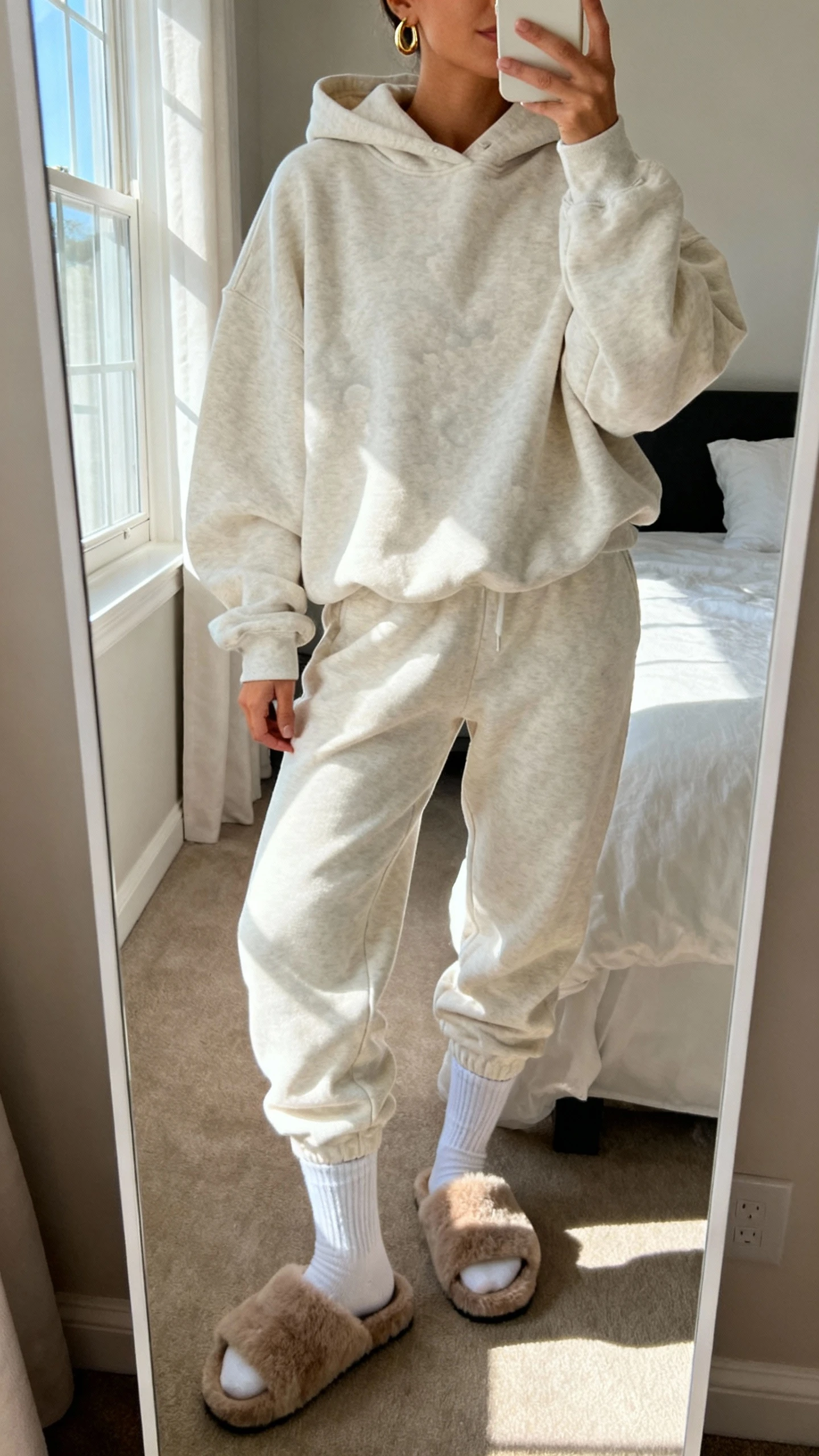 iPhone mirror selfie of a woman wearing a cloud-soft matching sweatsuit (oversized hoodie and joggers) with chunky socks, minimal gold hoops, and fuzzy house slides, face not visible, cropped at neck, bedroom mirror with morning window light, iPhone photo quality.