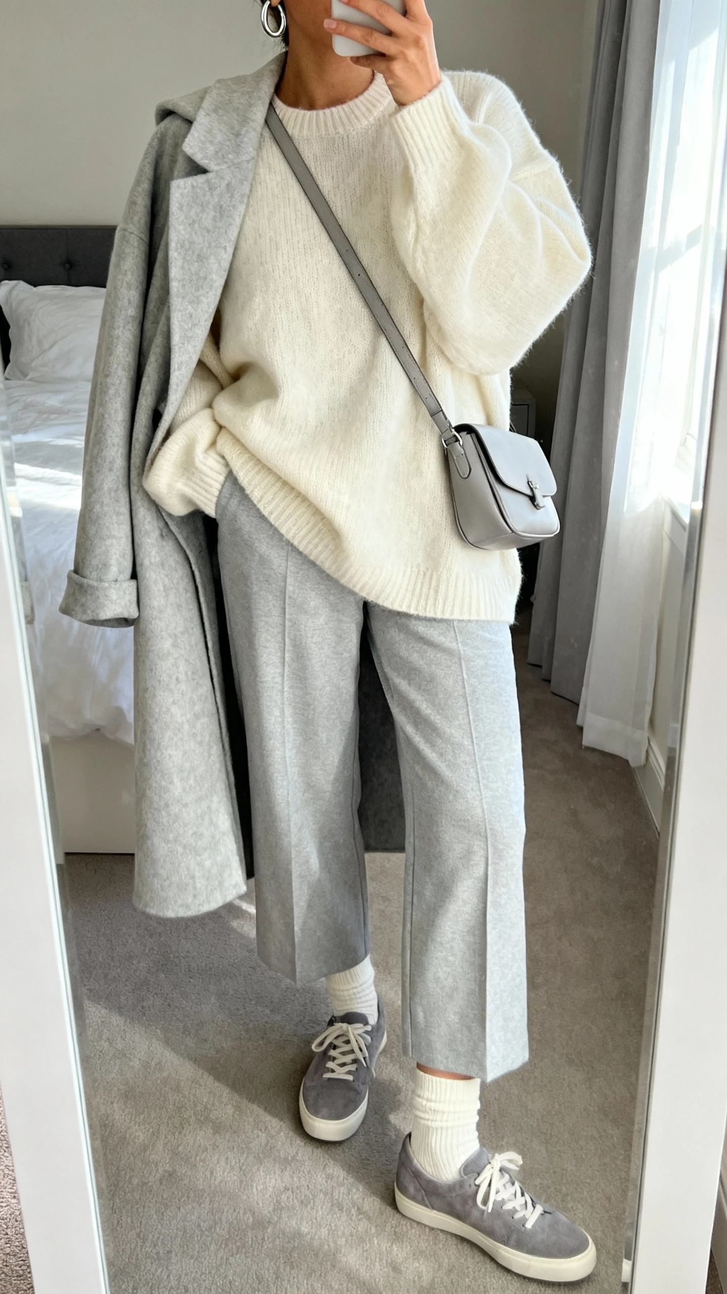 iPhone mirror selfie of a woman wearing a cloud-soft monochrome outfit: cream oversized knit sweater, light gray wide-leg trousers, pale gray wool coat draped over shoulders, cream socks and gray suede sneakers, silver minimalist hoop earrings, light gray leather crossbody; face not visible, cropped at neck, bedroom mirror, soft morning window light, iPhone photo quality.